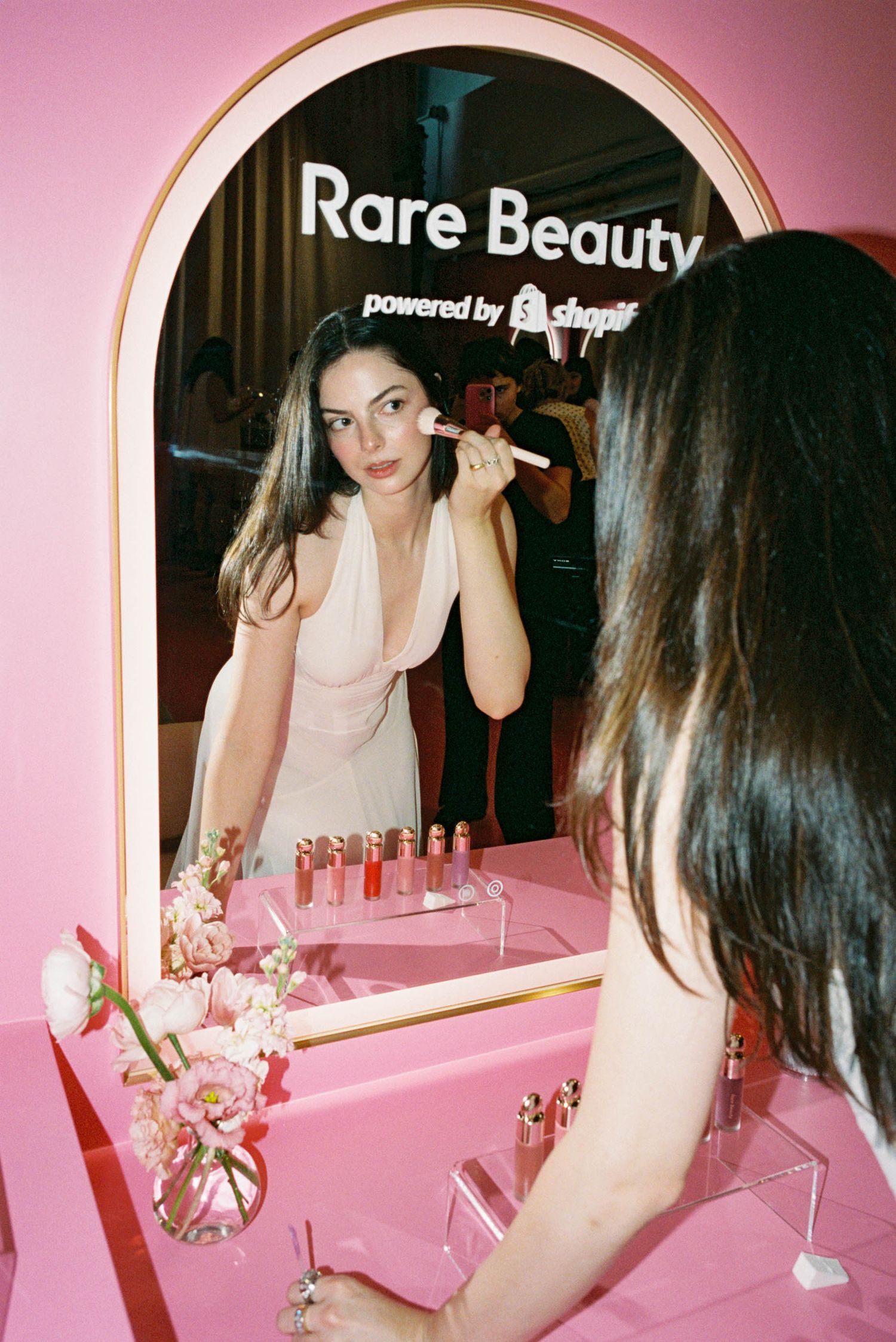 Pink vanity area with arched mirror displaying beauty products and fresh flowers.