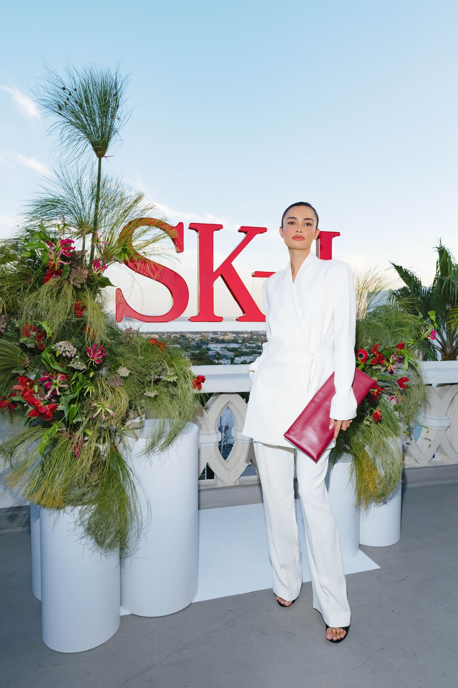 Person in all-white ensemble poses against SK-II branded backdrop with festive greenery.