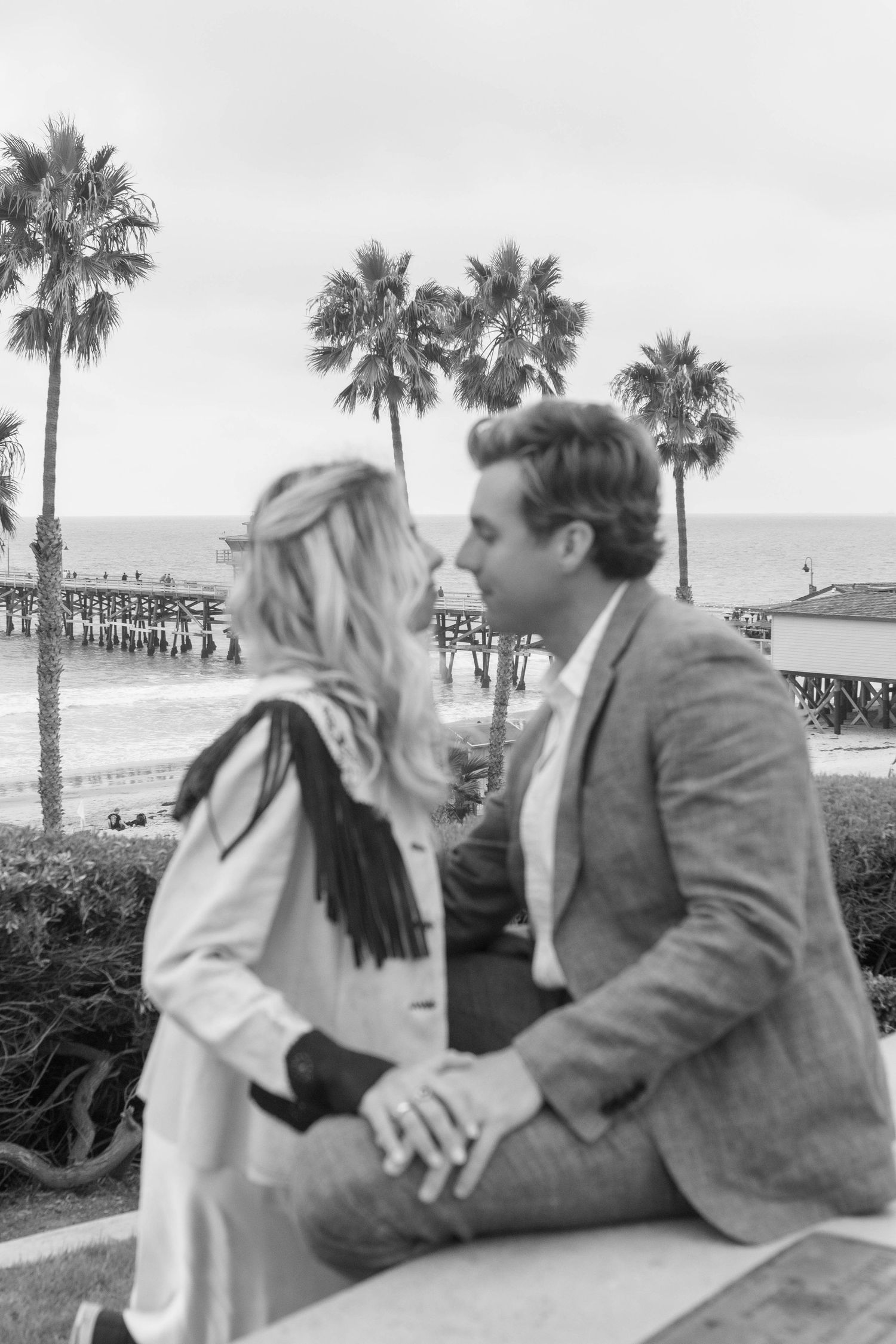 A couple sits close together on a bench overlooking a beach with palm trees in the background.