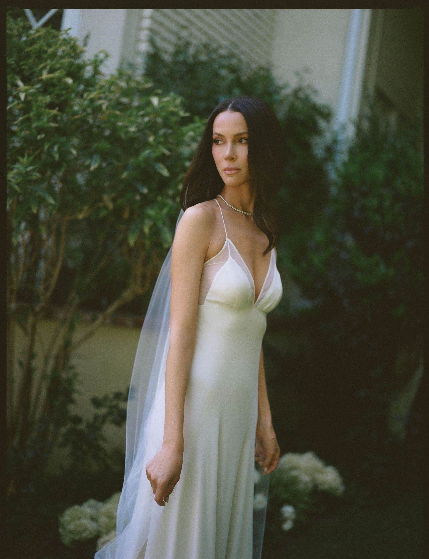 Woman in white satin slip wedding gown with veil poses outdoors among lush green garden foliage.