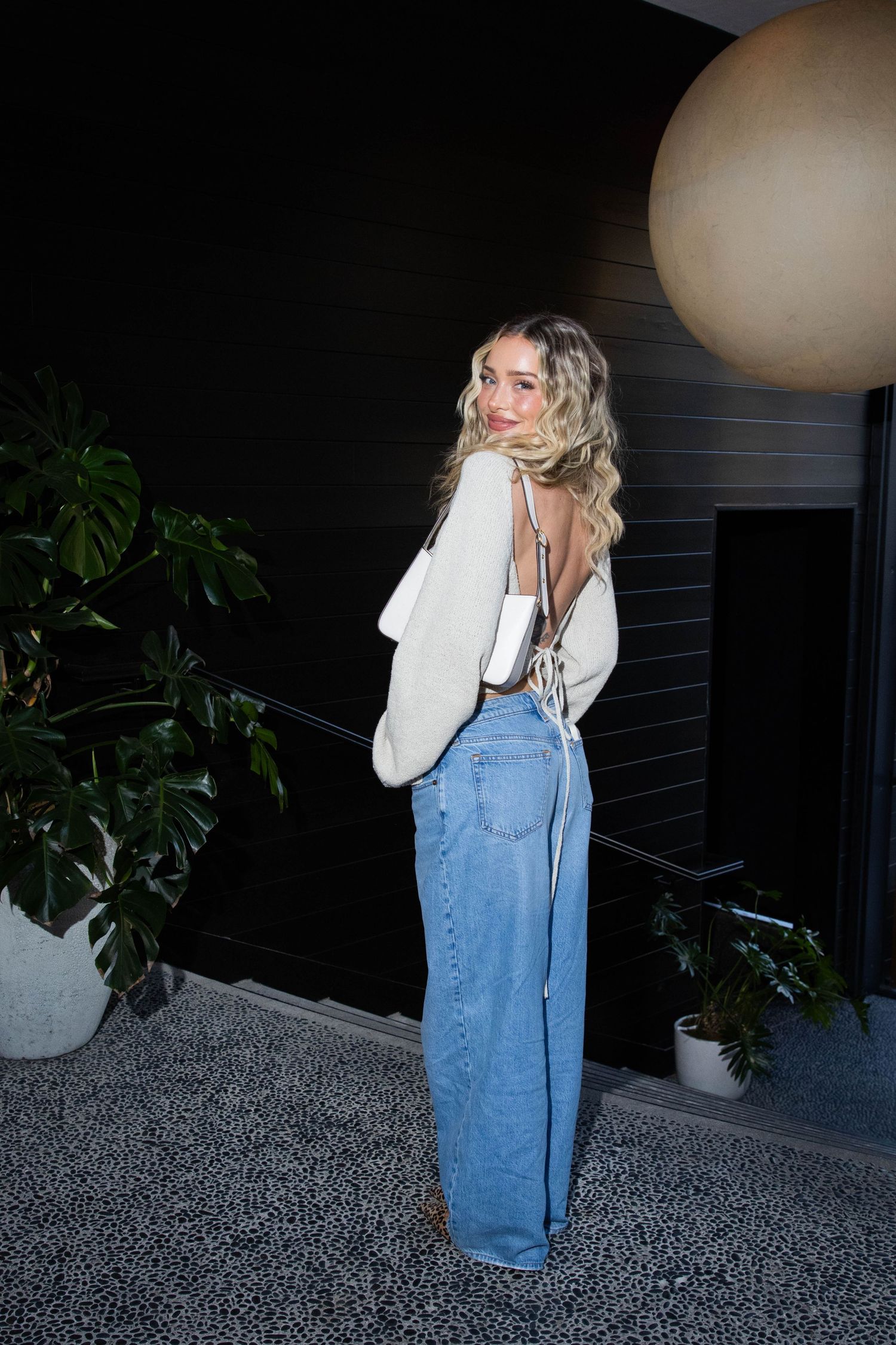Woman in white blazer and wide-leg jeans poses in front of dark wall with indoor plants and decorative sphere.