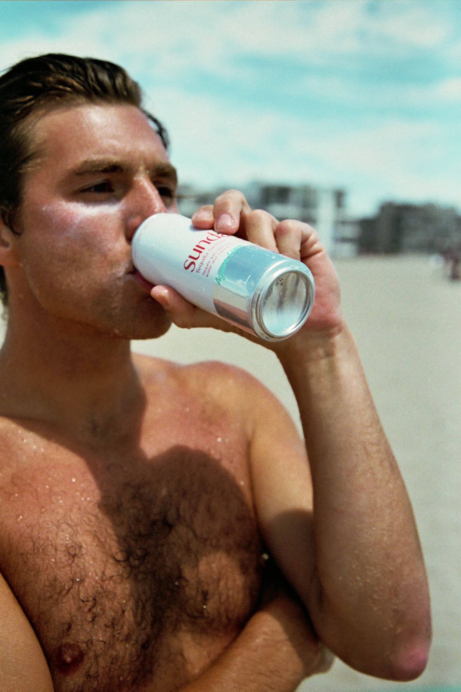 A shirtless beachgoer drinks from a white beverage can on a sunny beach.