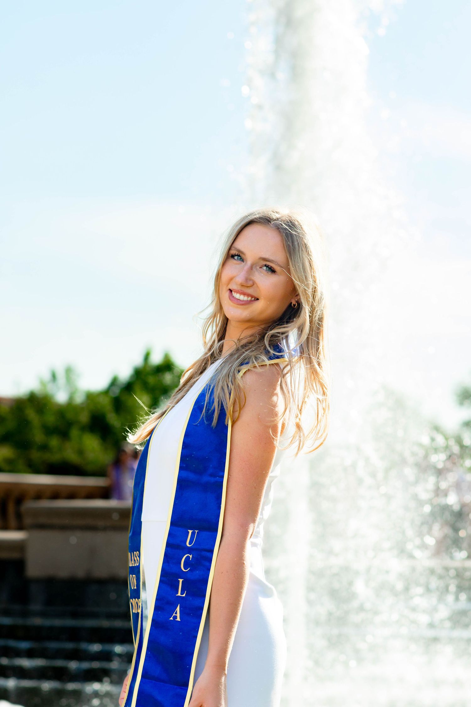 Graduate in white dress with blue stole poses by fountain during graduation photoshoot.