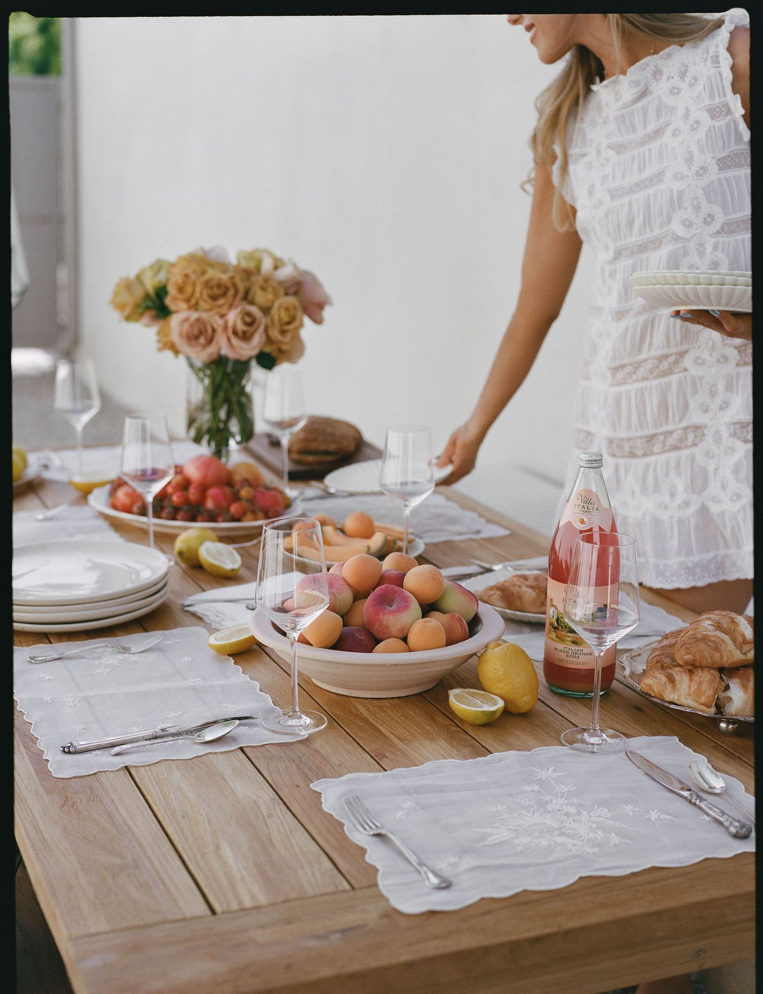 Woman in white dress setting a wooden table with fresh fruit, roses, croissants, and glassware for an elegant outdoor brunch.