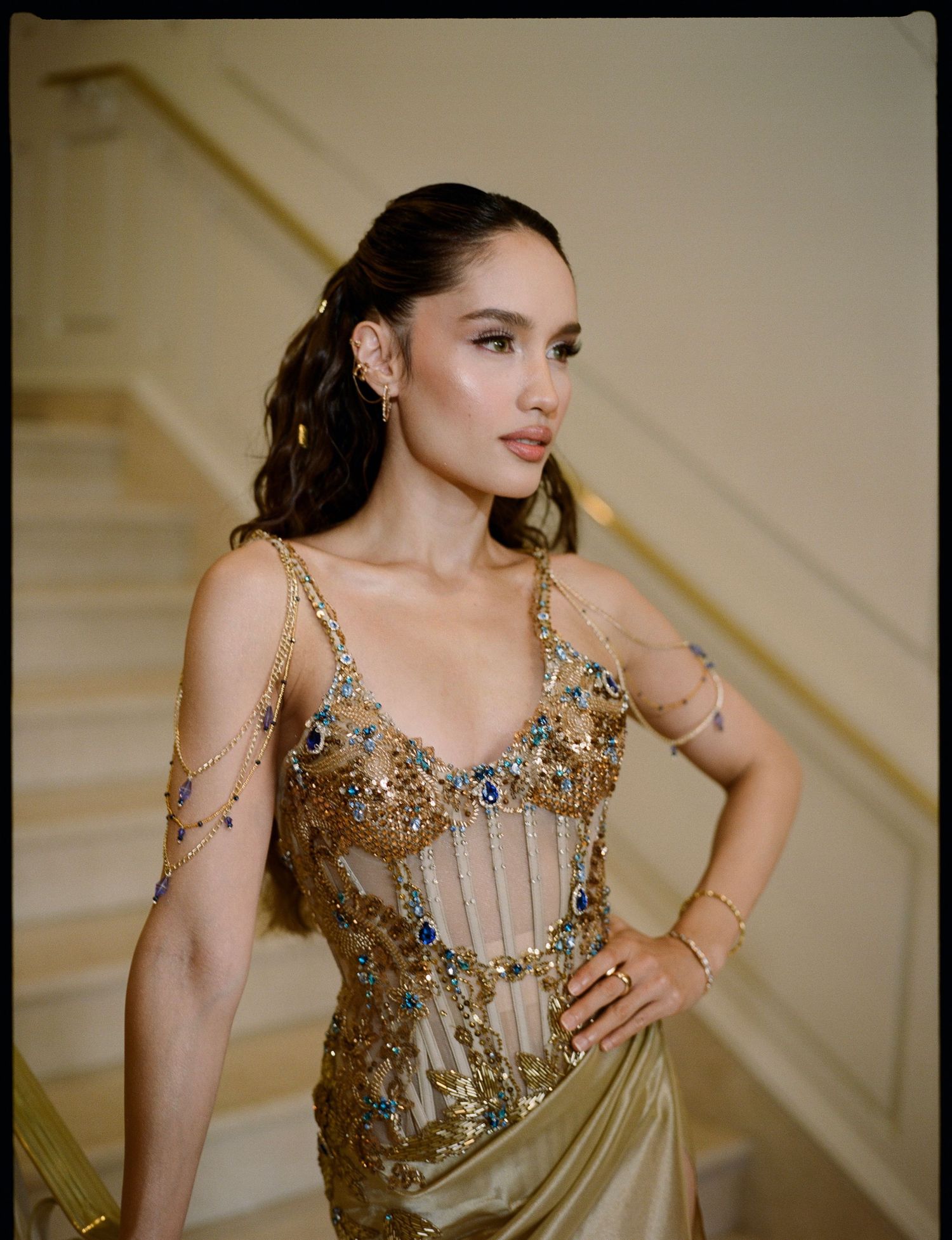 Woman with dark hair pulled back wears a gold jeweled corset gown with chain strap details while posing beside an elegant staircase.