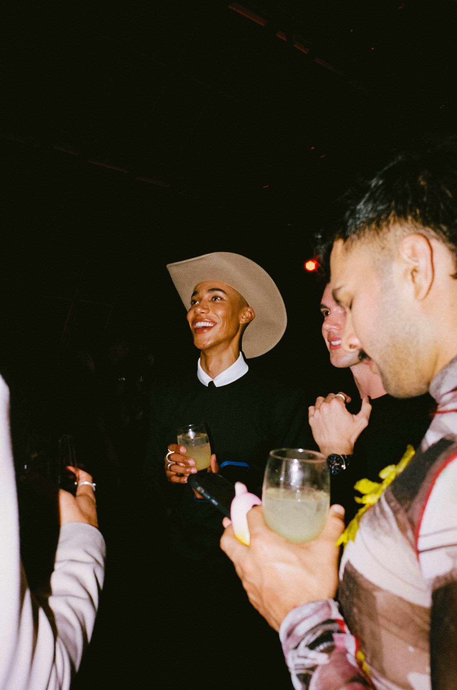 People socializing at a party, one wearing a wide-brim hat, holding cocktails in a dimly lit venue.