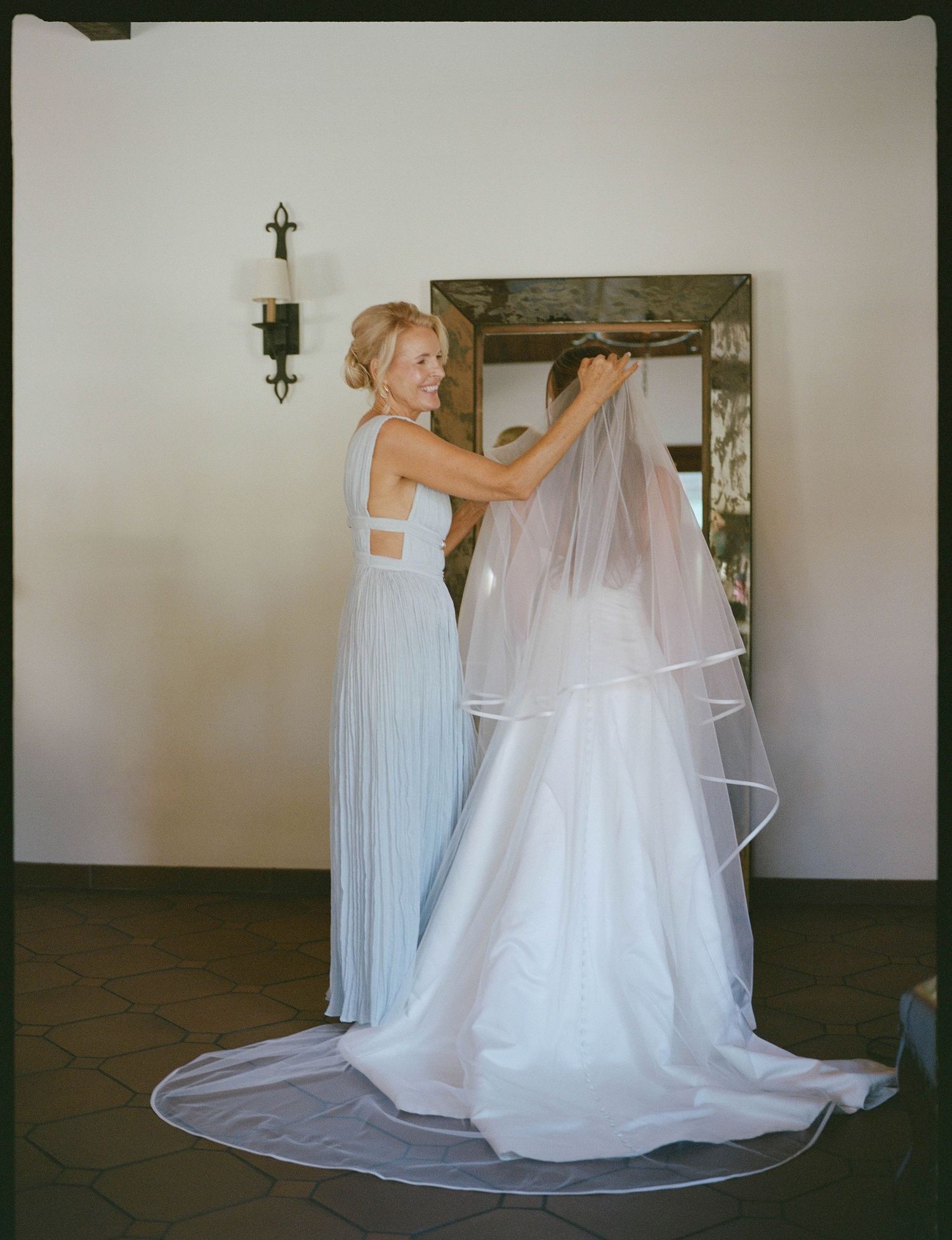 A smiling woman in a pale blue gown adjusts the long tulle veil of a bride in a white wedding dress in front of a mirror.