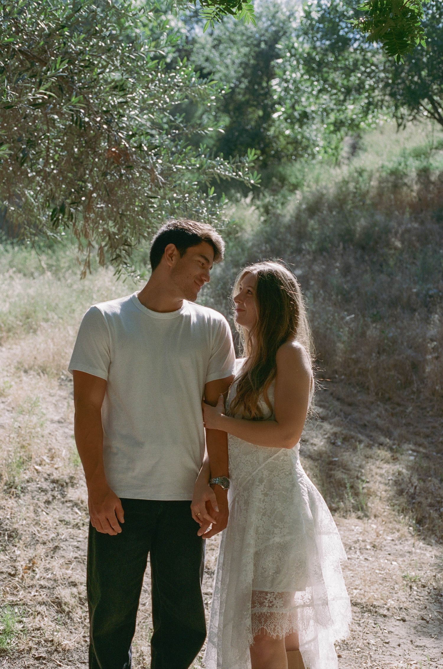 A couple shares a romantic moment on a sunlit nature trail, the woman in a white lace dress smiling up at the man in a white tee.