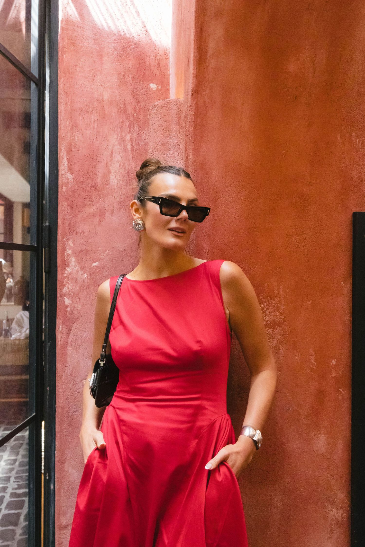 Woman in vibrant red sleeveless dress poses against a rustic terracotta wall.