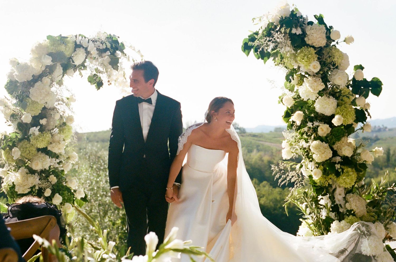 Wedding couple standing under floral arch with white roses against scenic backdrop.