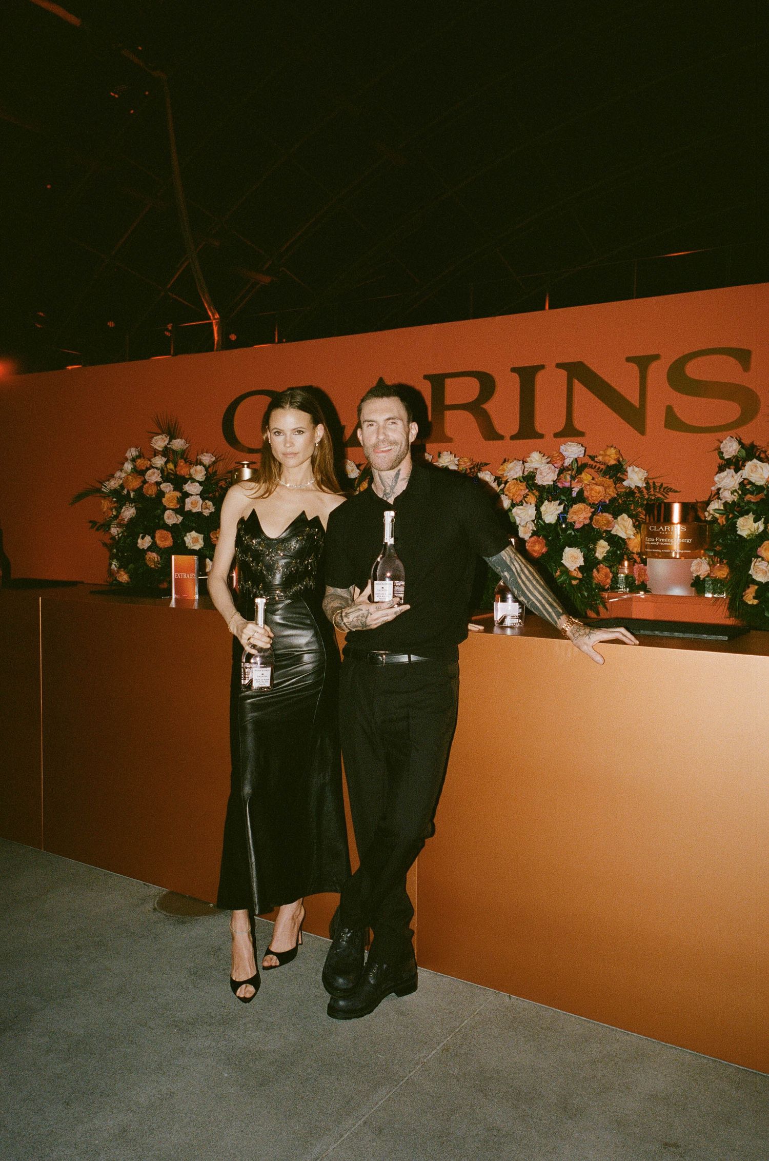 Stylish duo holds bottles at Clarins branded display with floral arrangements at upscale event.