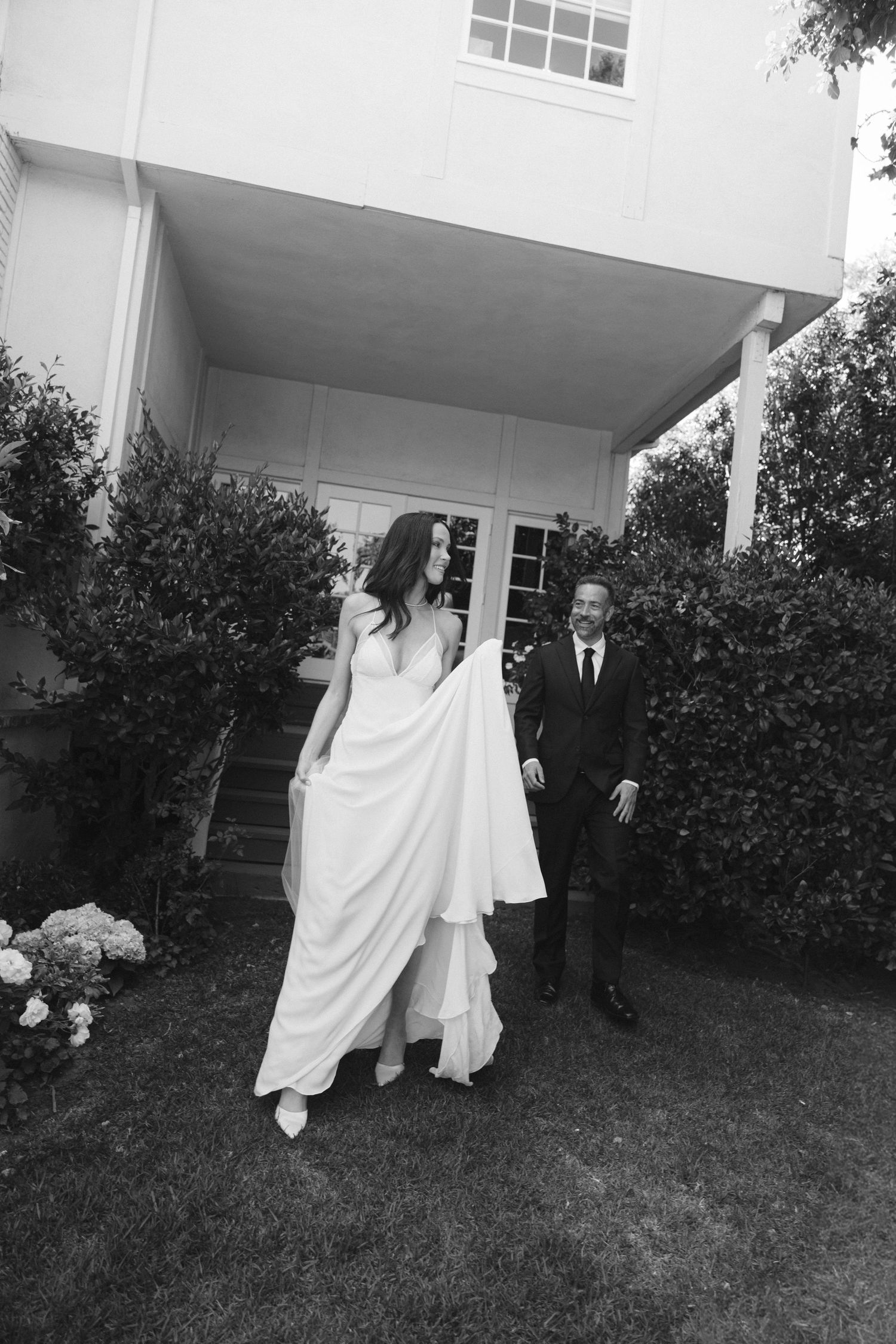 Black and white photo of a bride in a flowing gown and groom in a suit walking outside a white house.