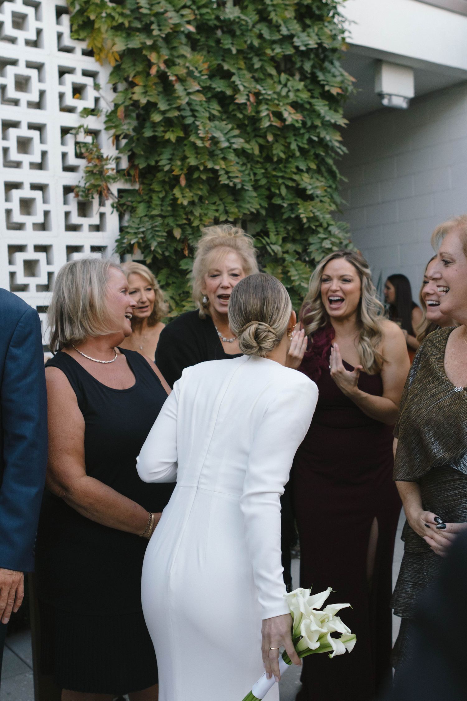 A group of people laughing together at an outdoor event with white lattice walls and green foliage in the background.