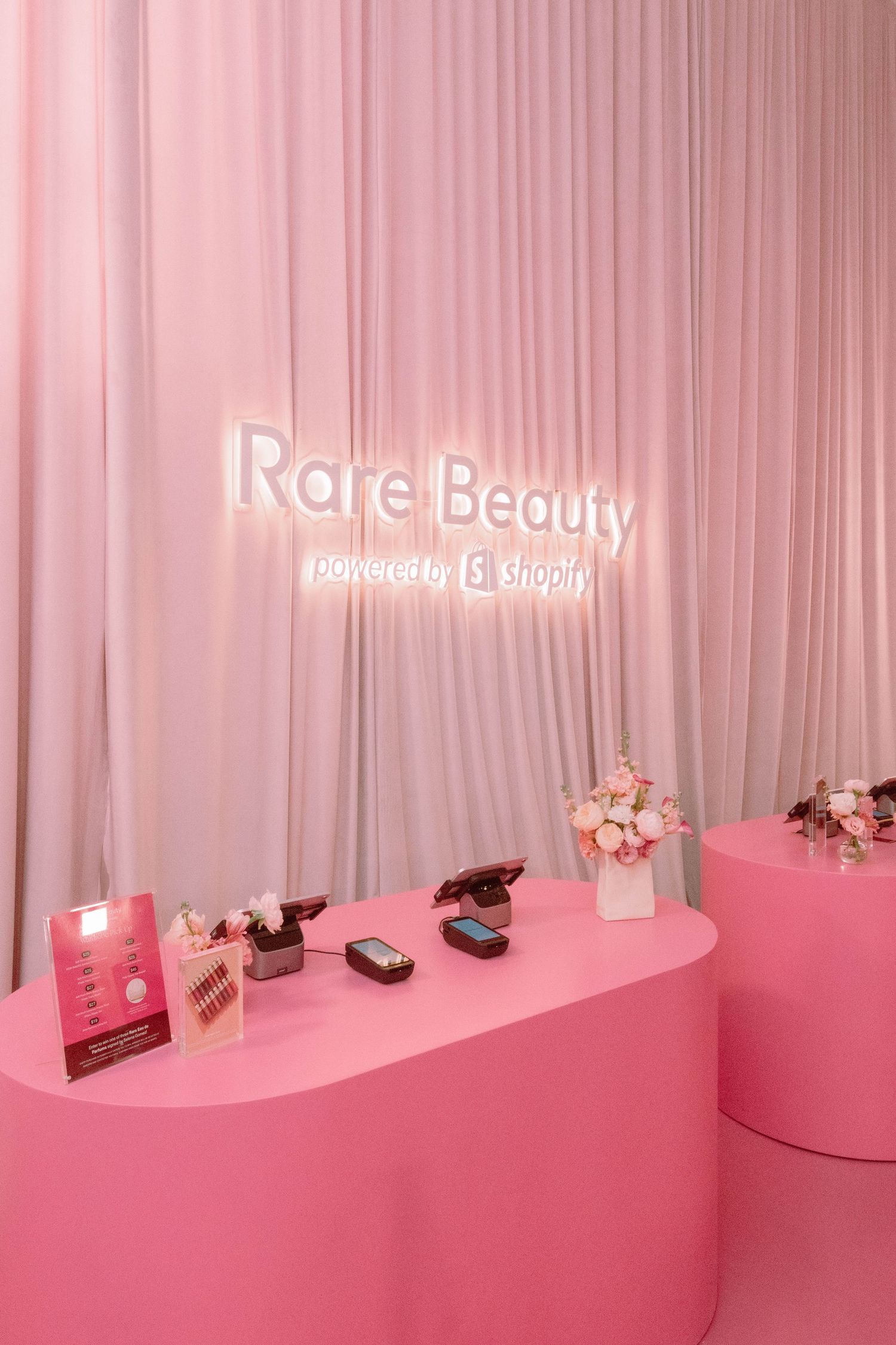 Pink-themed retail display with beauty products arranged on curved white shelving against a draped backdrop.