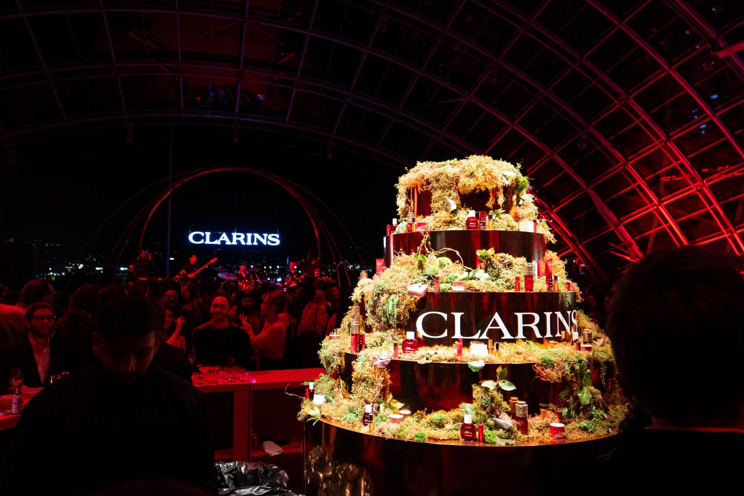Elaborate tiered cake with floral decorations displayed at Clarins event under red lighting.
