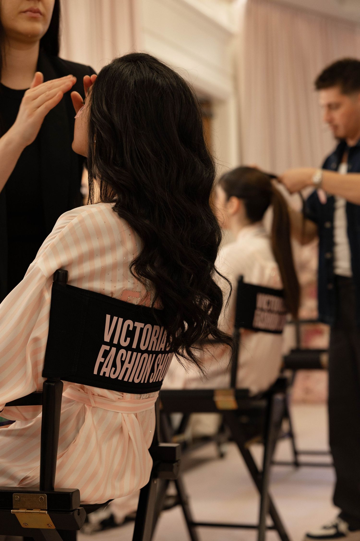 Backstage at the Victoria's Fashion Show, a model with long dark wavy hair sits in a director's chair while stylists prepare her look.