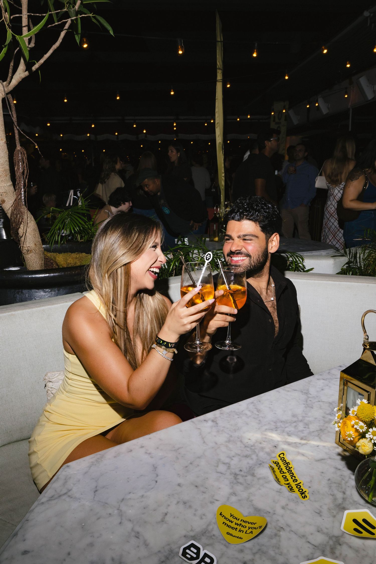 A couple smiles and clinks cocktail glasses at a marble table in a lively outdoor bar at night.