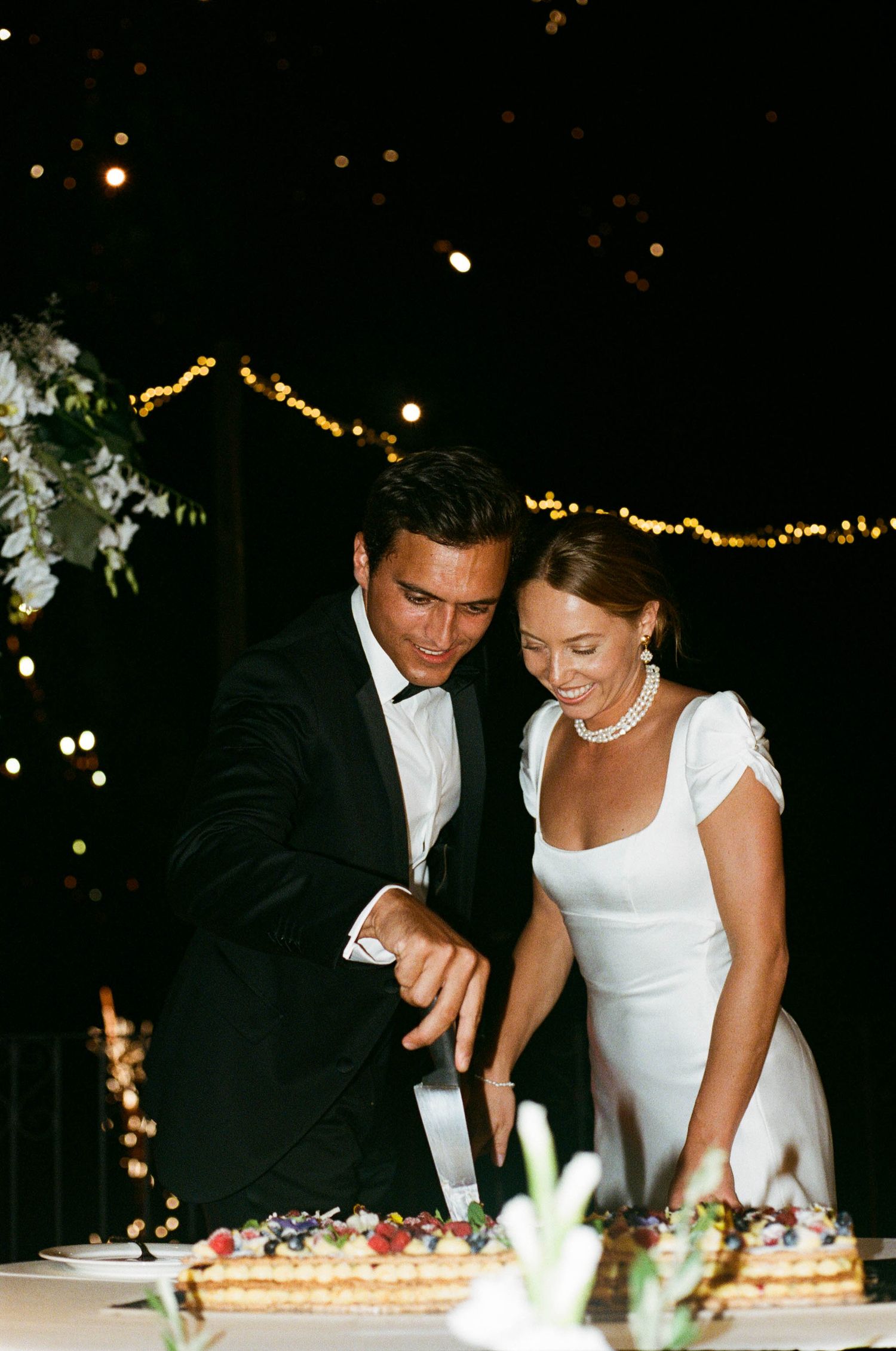 A couple in formal attire cuts their wedding cake together at an evening reception.