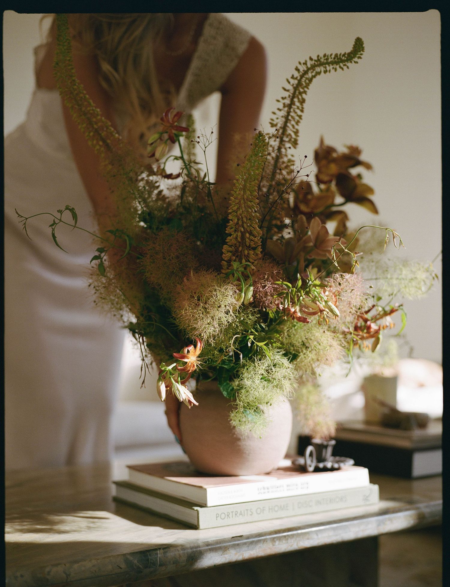 A lush floral arrangement in a round terracotta vase sits atop stacked books on a wooden table, with a woman in white dress behind.