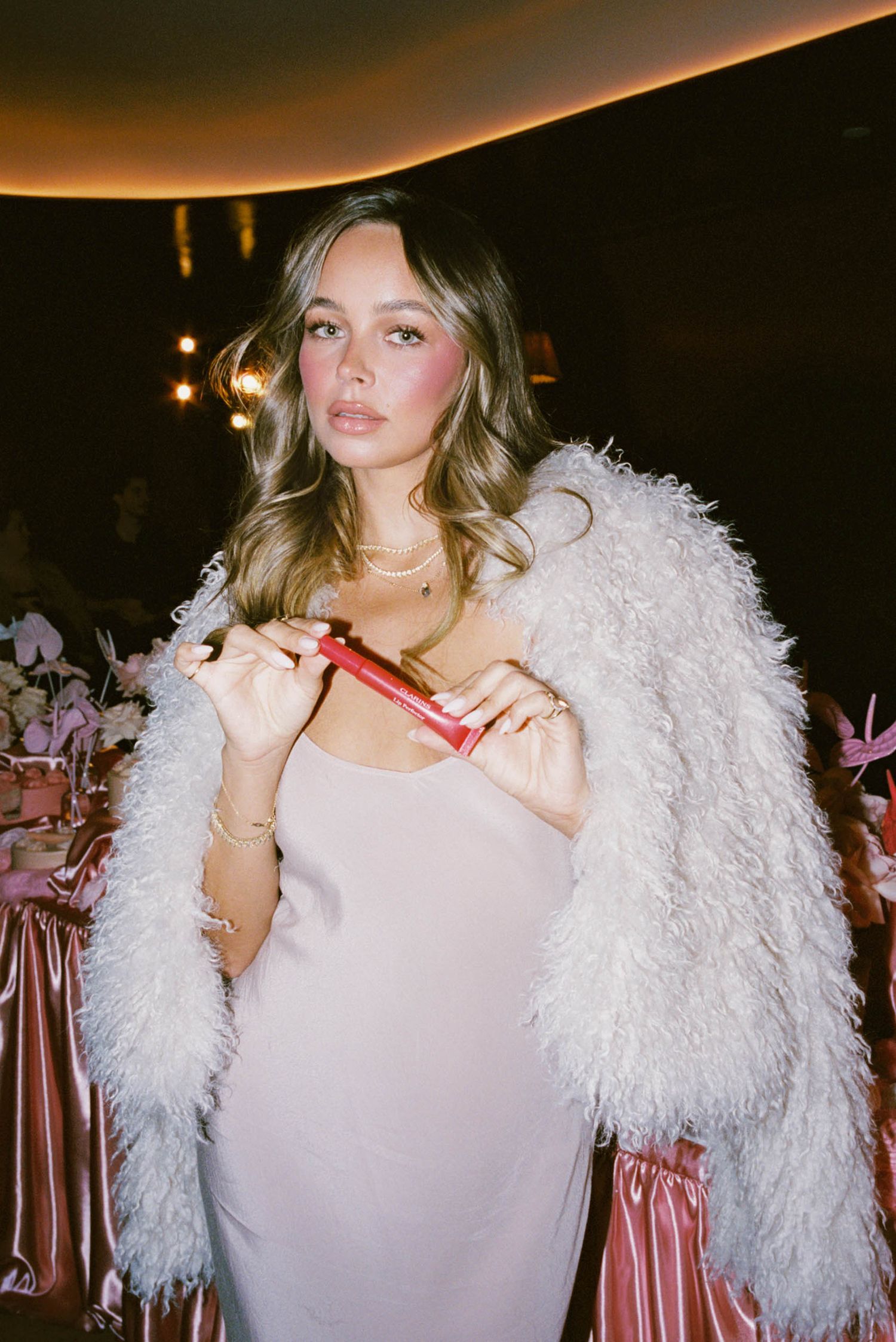 A person in a white fluffy jacket poses playfully in a dimly lit room with warm ambient lighting.