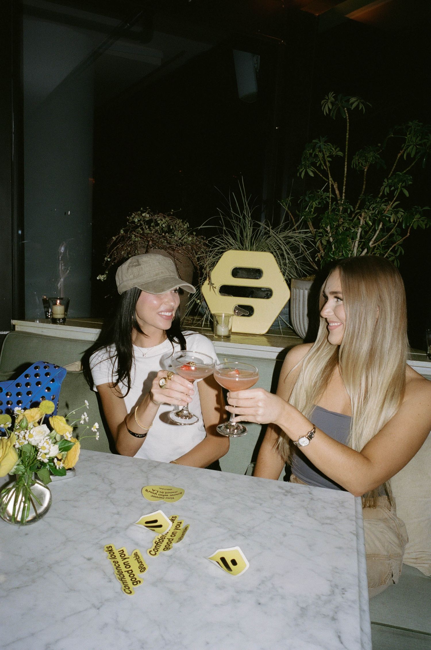 Two women smiling and clinking cocktail glasses at a marble table in a stylish bar setting.