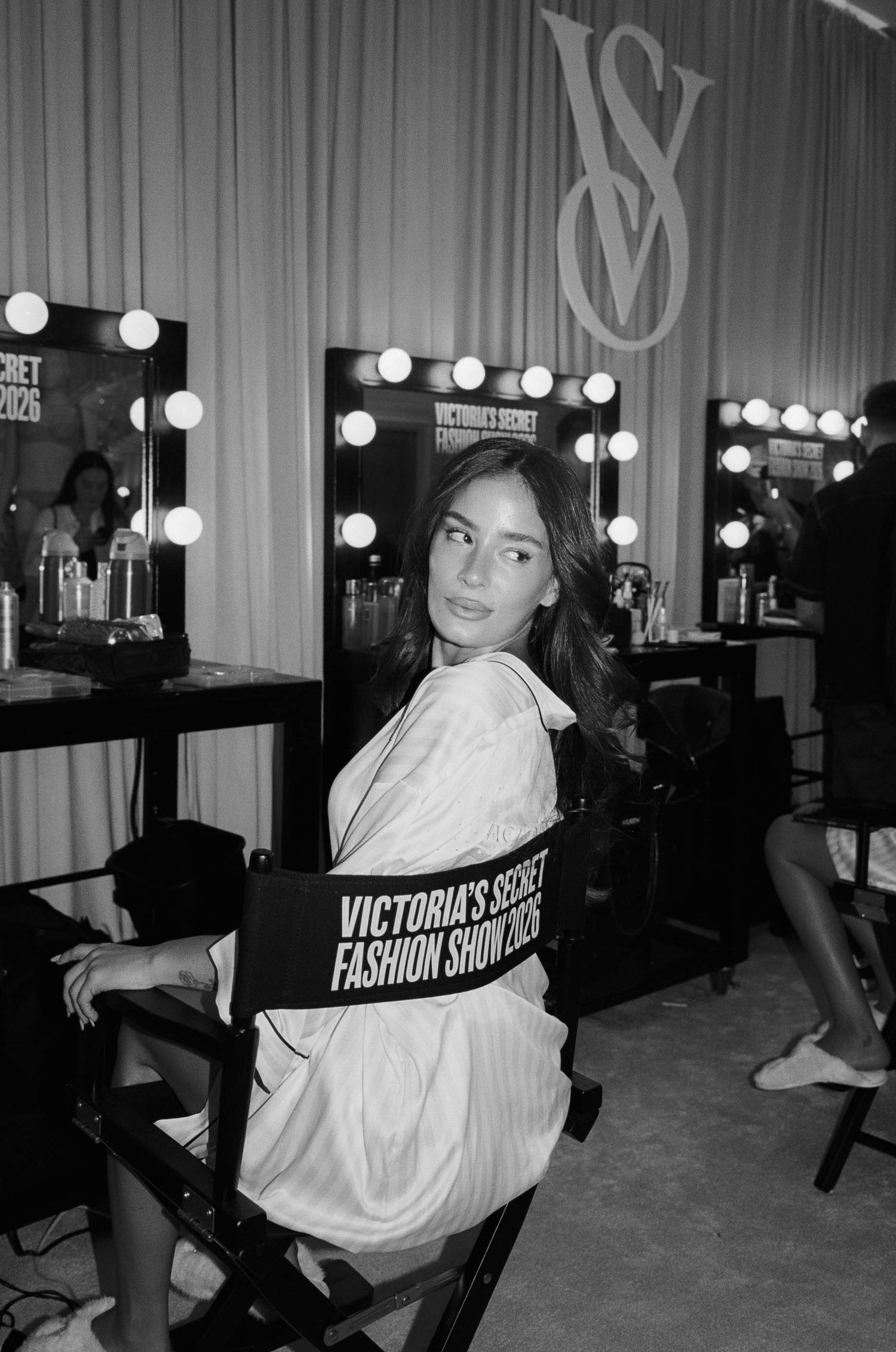Woman in a satin robe sits in a Victoria's Secret Fashion Show director's chair backstage beside lighted vanity mirrors in black and white.