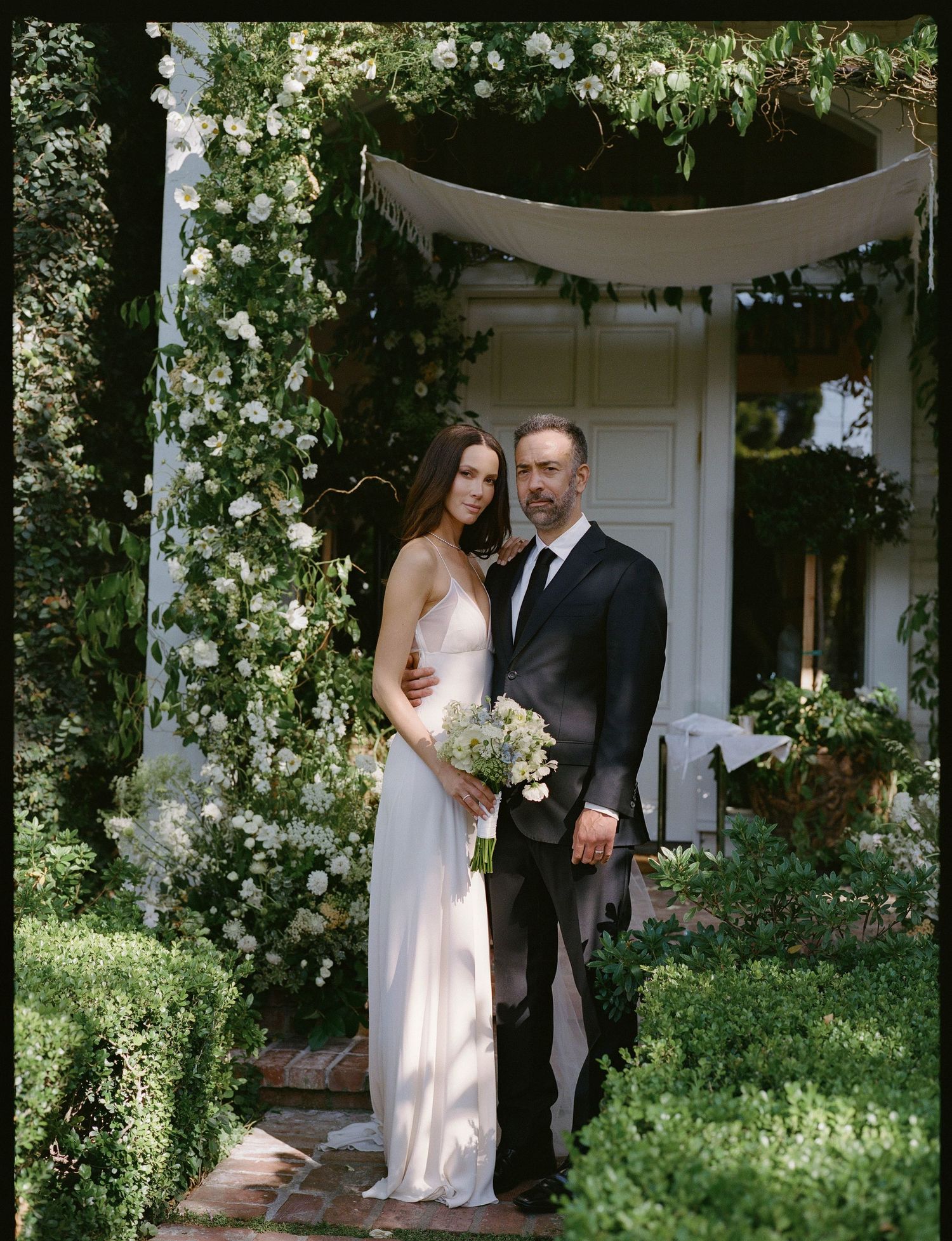 Couple poses under floral arch at outdoor wedding ceremony, bride in white gown holding bouquet, groom in dark suit.