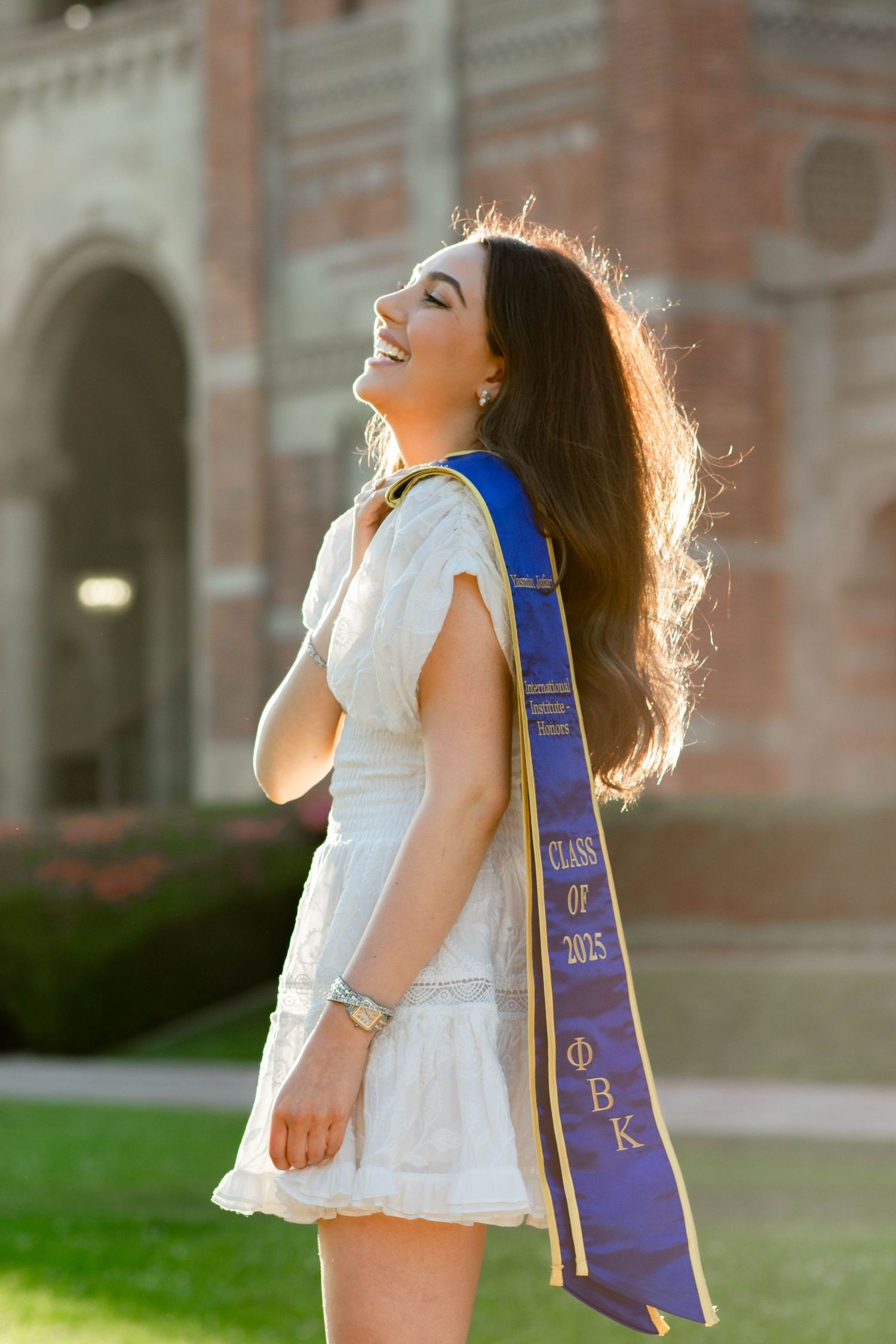 Graduate in white dress and purple stole dances joyfully in golden sunlight on campus lawn.