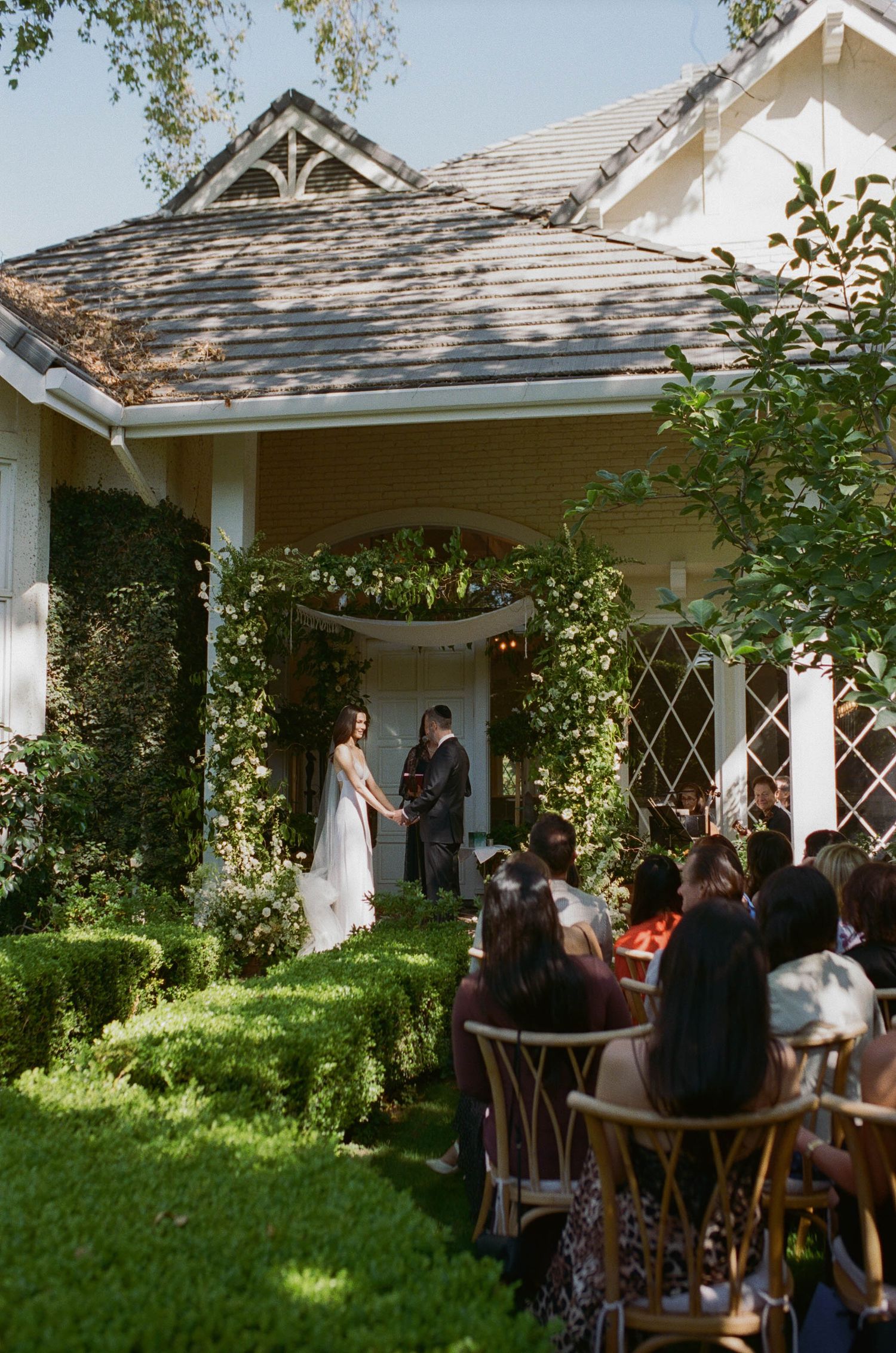 Outdoor wedding ceremony at a charming cottage venue with guests seated on a sunny garden lawn.