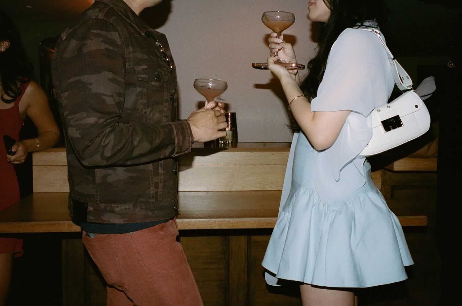 Two people holding cocktail glasses at a party, one wearing a light blue dress with a white bag.