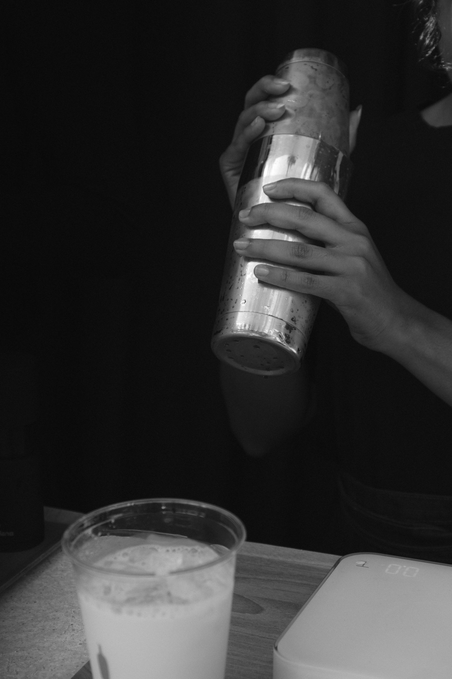 Black and white photo of bartender shaking cocktail shaker over frothy drink in plastic cup on wooden bar.