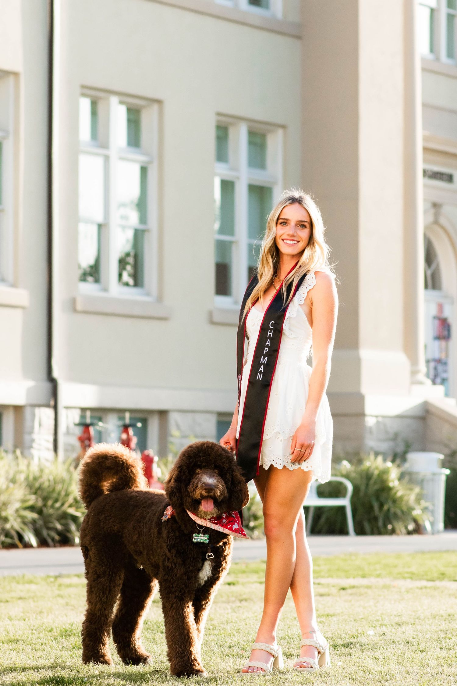 Graduate in white dress poses with black dog on campus lawn.