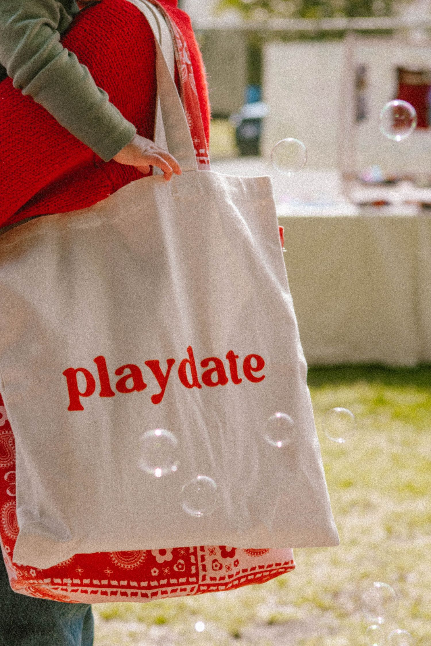 White canvas tote bag with red Playdate logo with soap bubbles floating in the background.