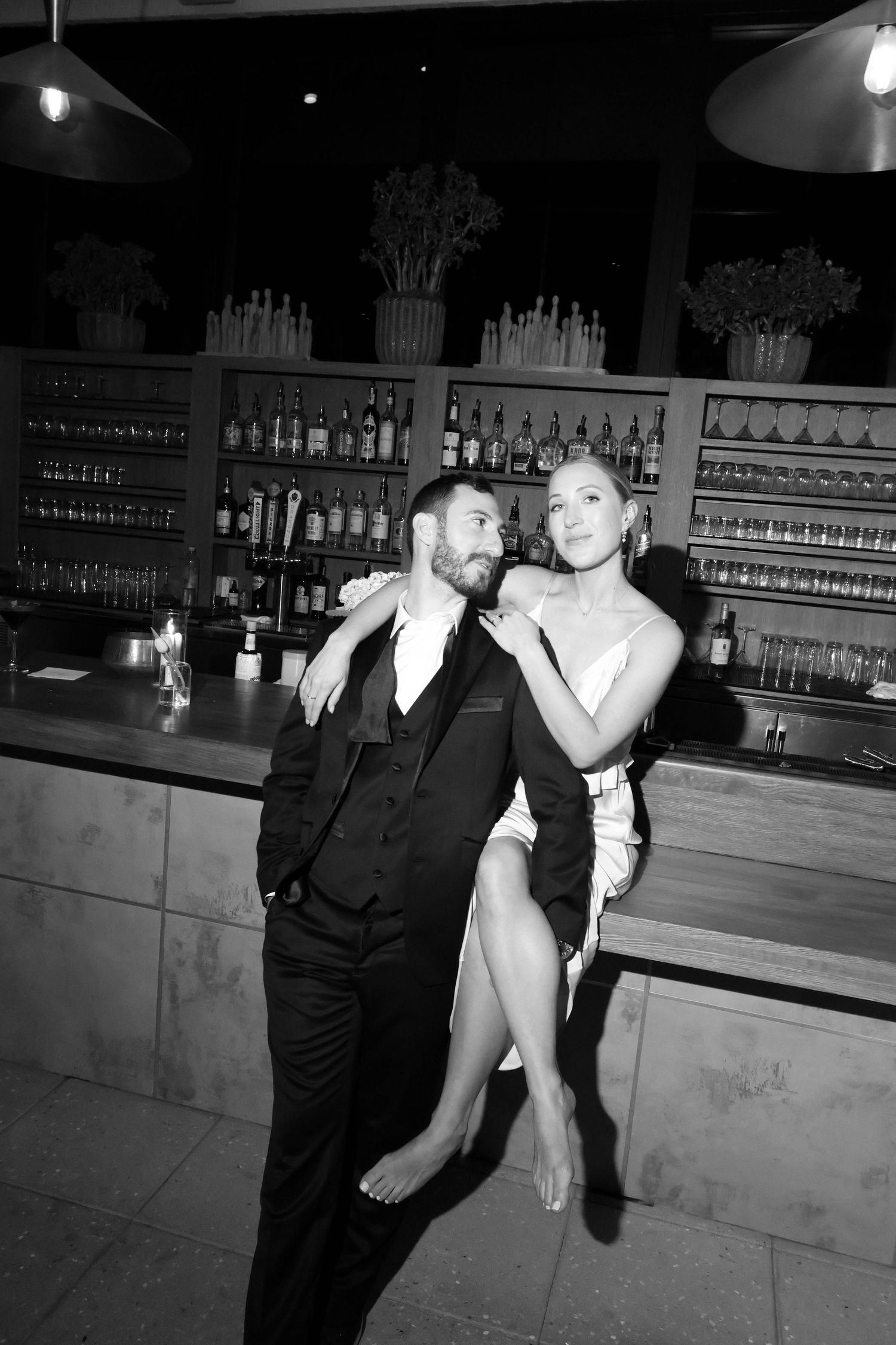 People in formal attire posing at a bar with bottles displayed in the background.