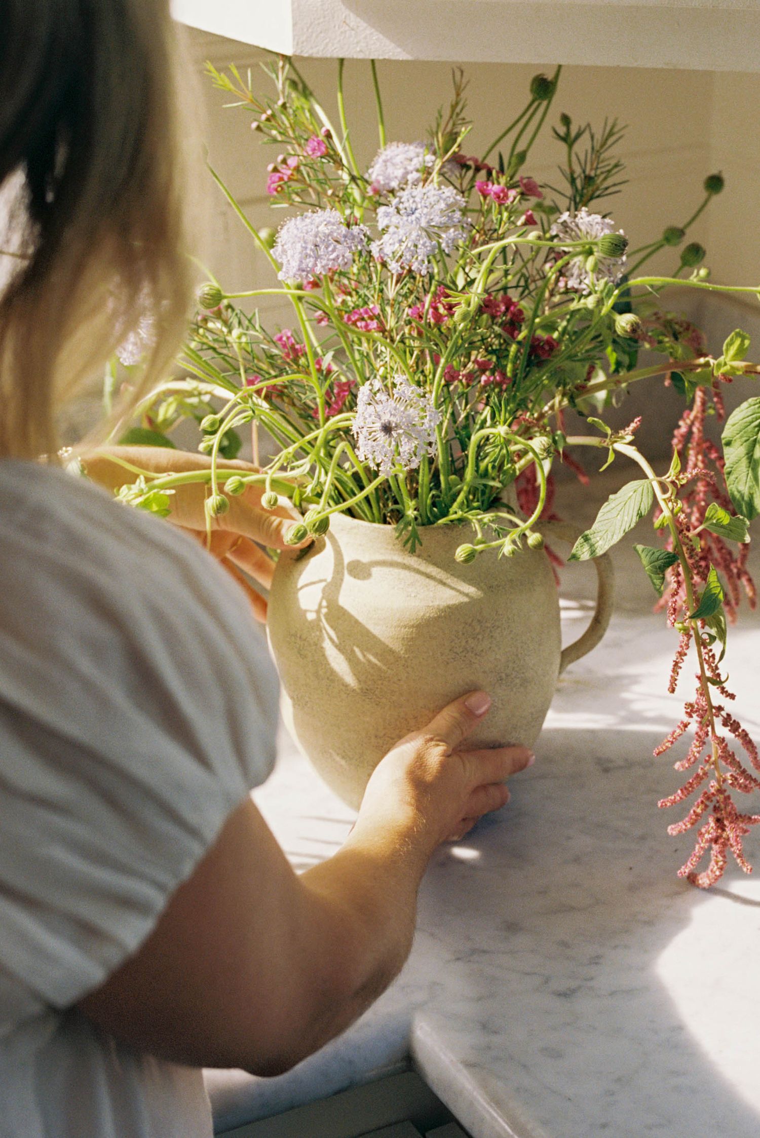 Hands arranging fresh flowers and greenery in a white vase on a marble countertop in natural sunlight.