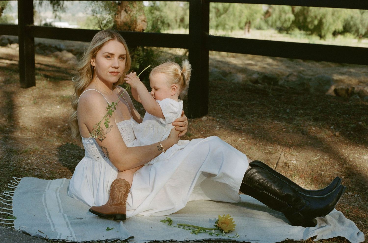 Young mother in white dress and cowboy boots sits on blanket outdoors with her toddler child.