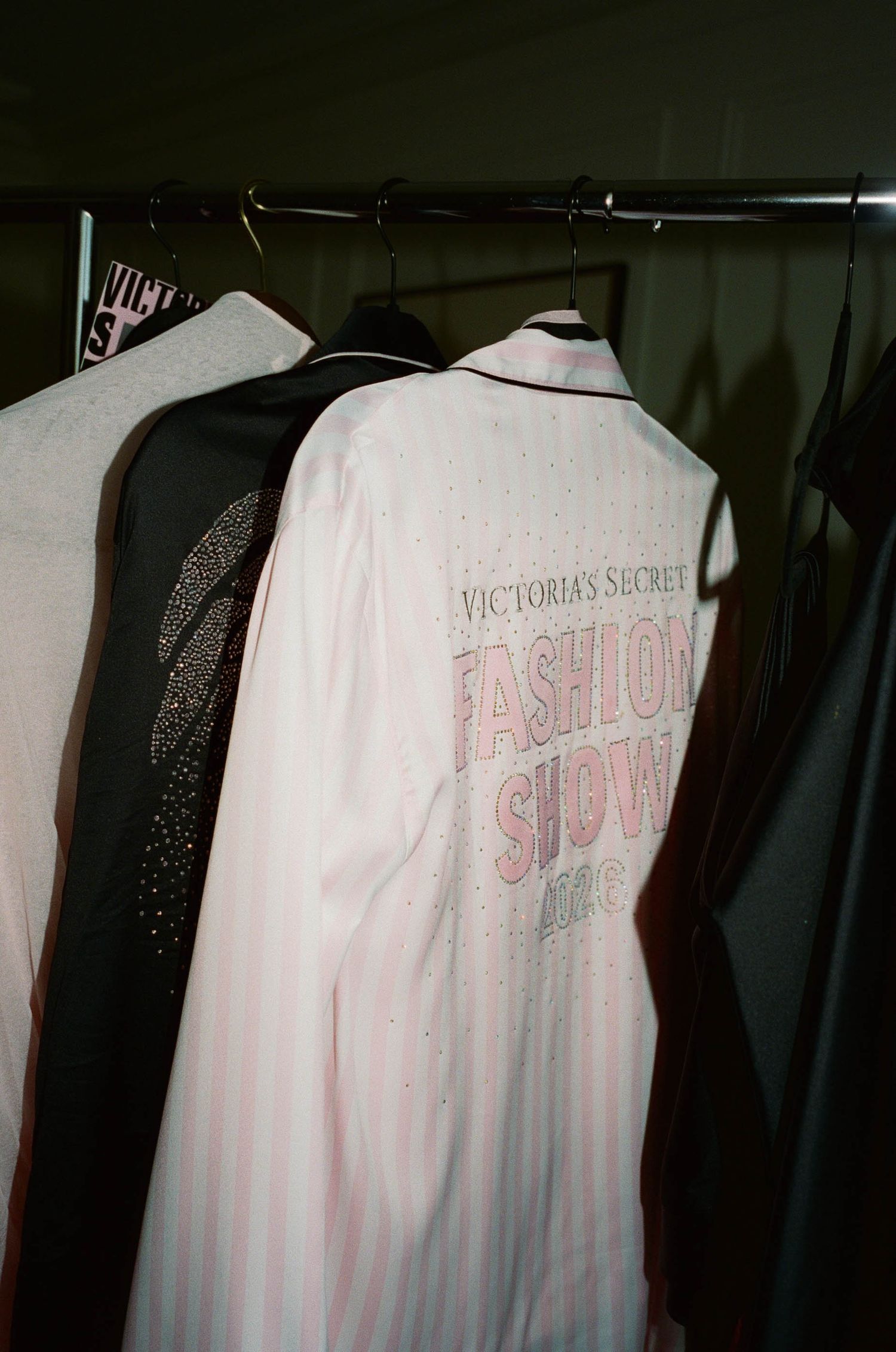 Pink striped satin pajama shirt printed with Victoria's Secret Fashion Show NYC hangs on a rack beside embellished jackets backstage.