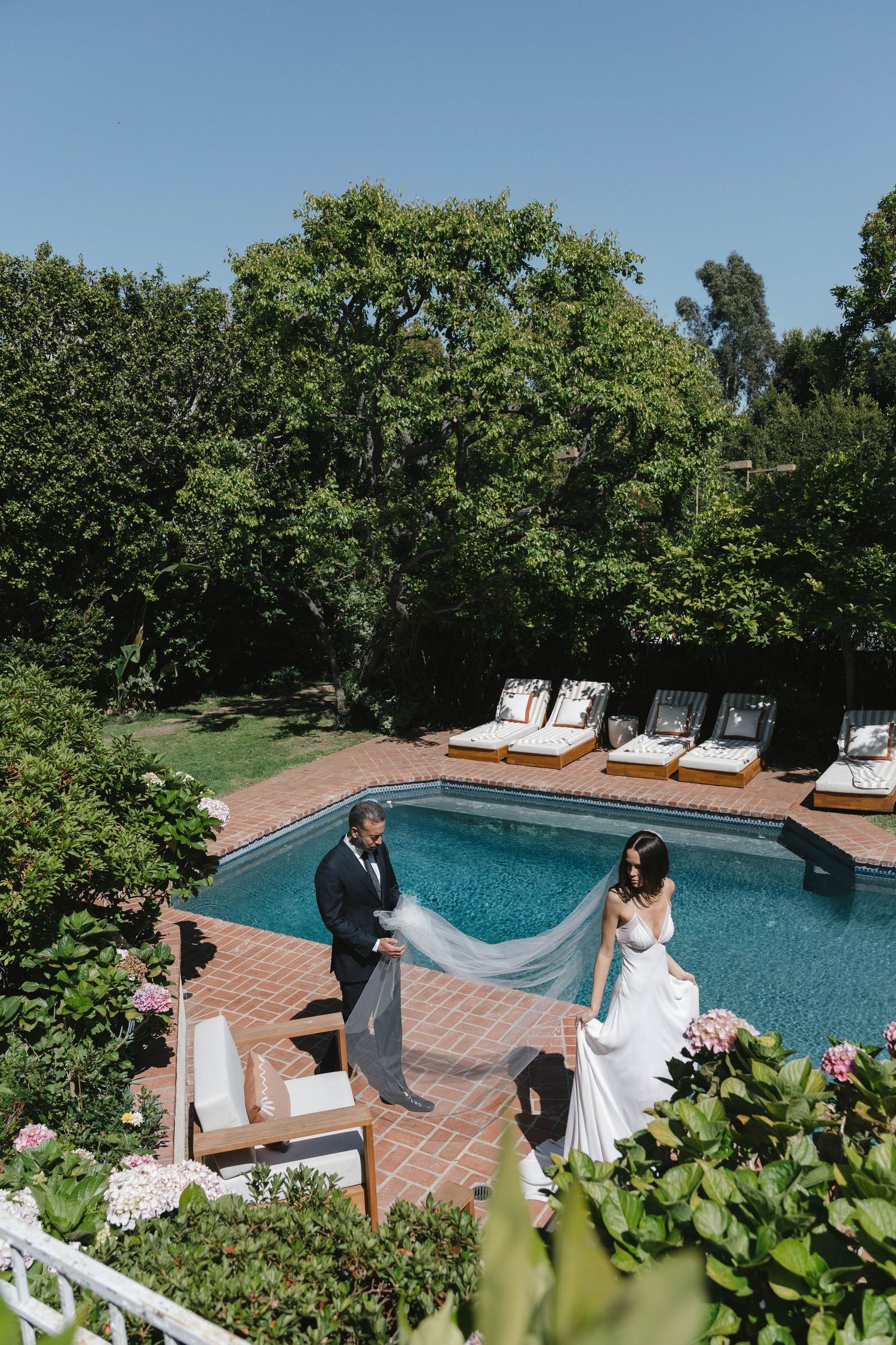 Bride in white gown and groom in suit pose by a luxury outdoor pool surrounded by lush greenery on a sunny day.