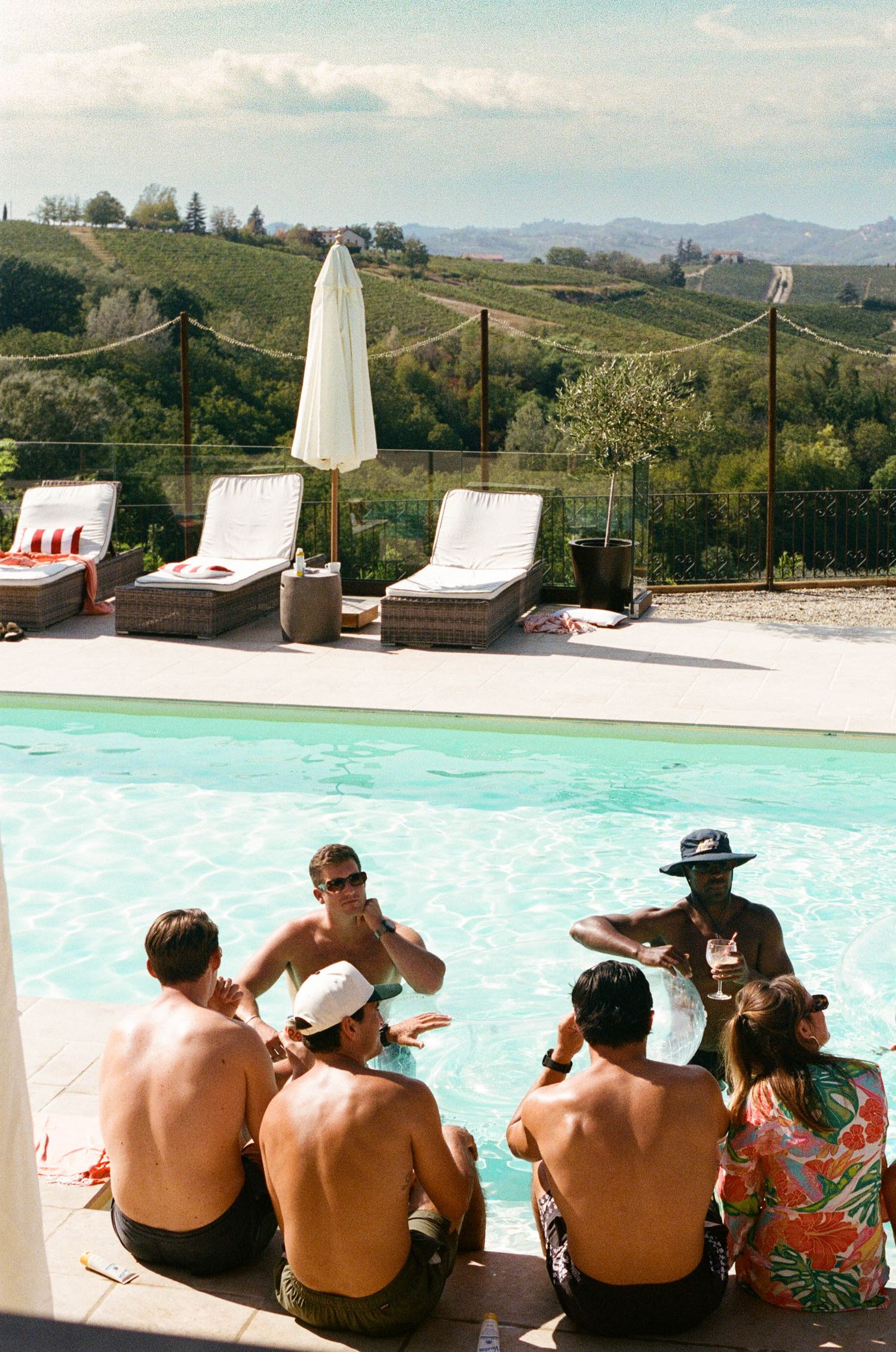 Group of people relaxing poolside with scenic vineyard hills in the background.