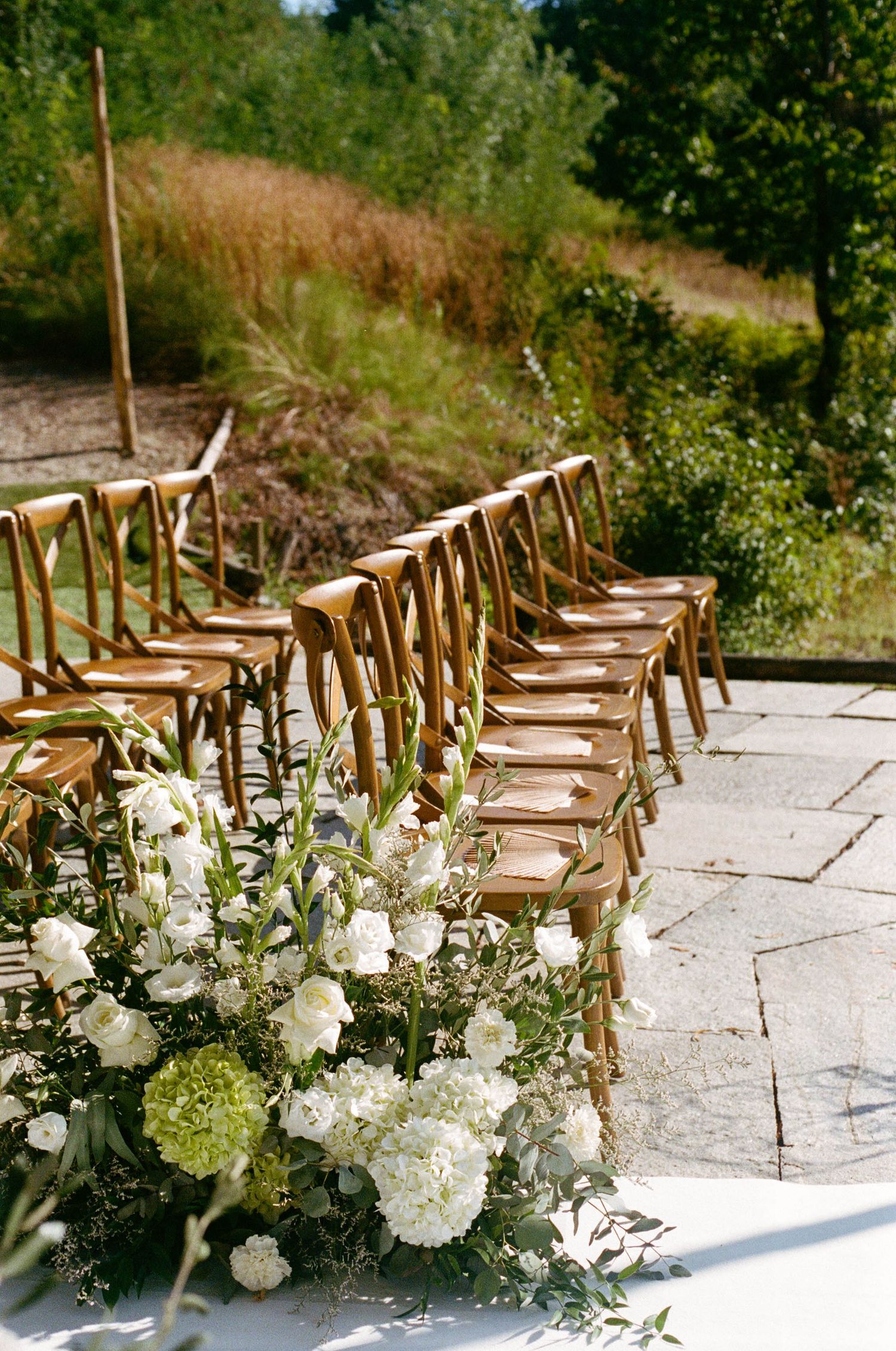 Wooden chairs lined up for outdoor wedding ceremony with white floral arrangements.