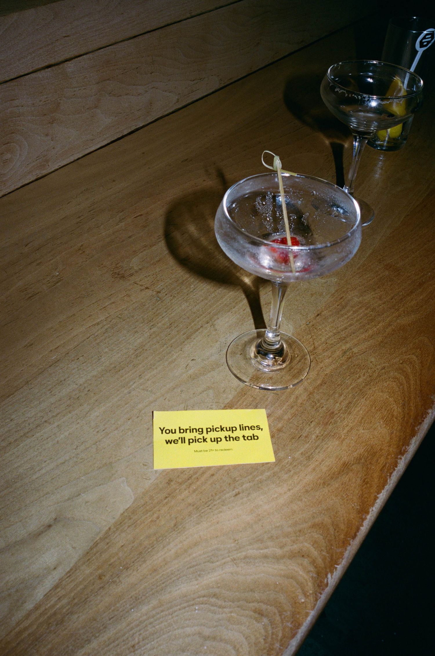 A cocktail glass with a cherry garnish on a wooden bar next to a yellow card reading You bring pickup lines, we'll pick up the tab.