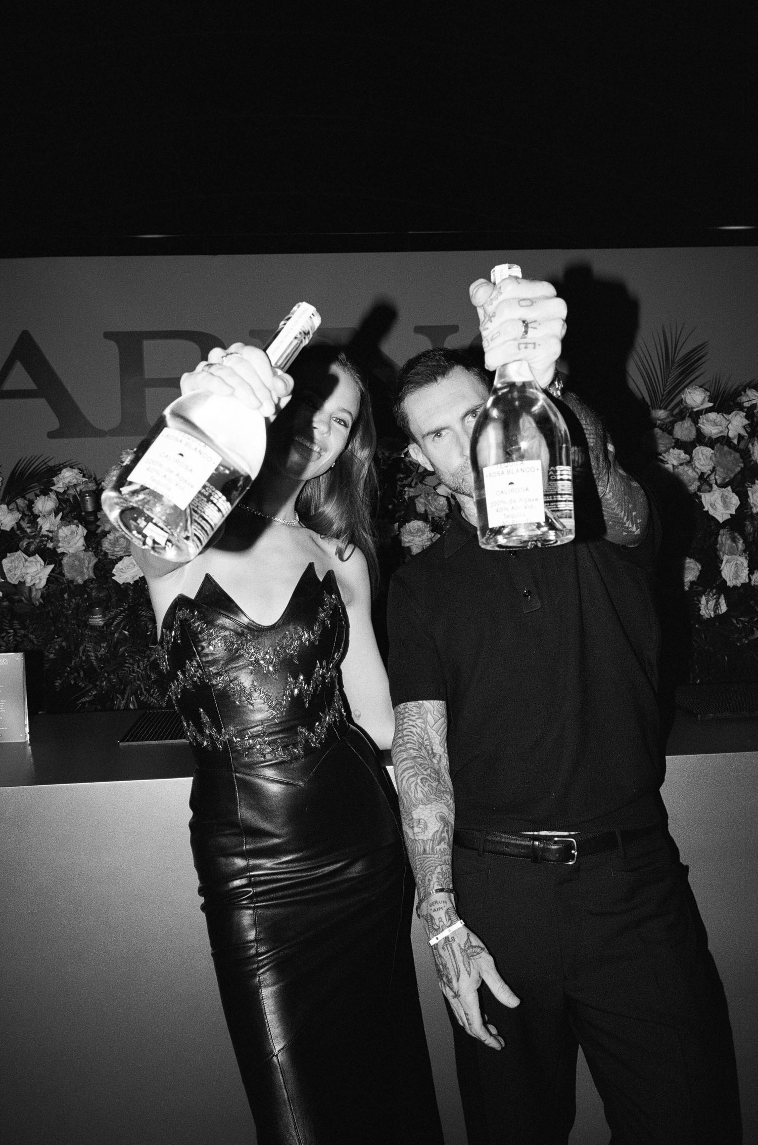 Black and white photo of tattooed couple raising bottles at Clarins party with flowers backdrop.