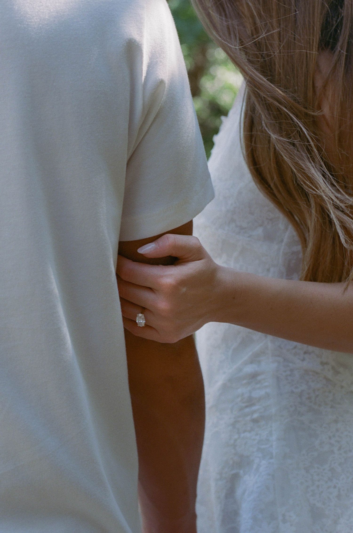 A bride in white lace holds her groom's waist, showcasing a sparkling diamond engagement ring.
