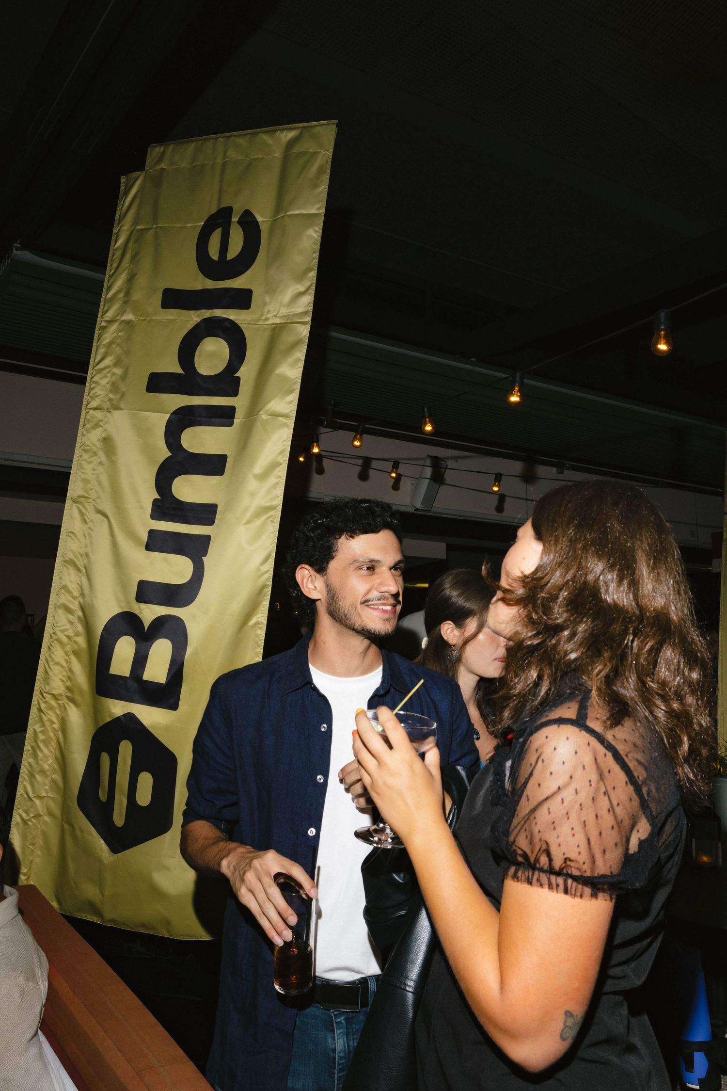 Man and woman socializing at a Bumble event, holding drinks near a yellow Bumble banner indoors.