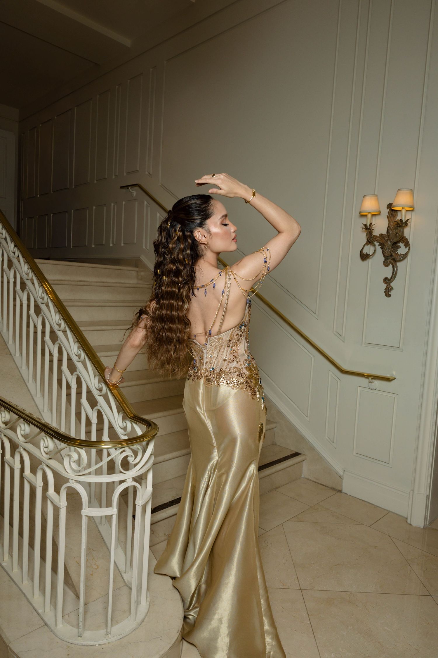 Woman in a gold satin gown with an embellished open back and long wavy hair poses with raised arms on an elegant staircase.