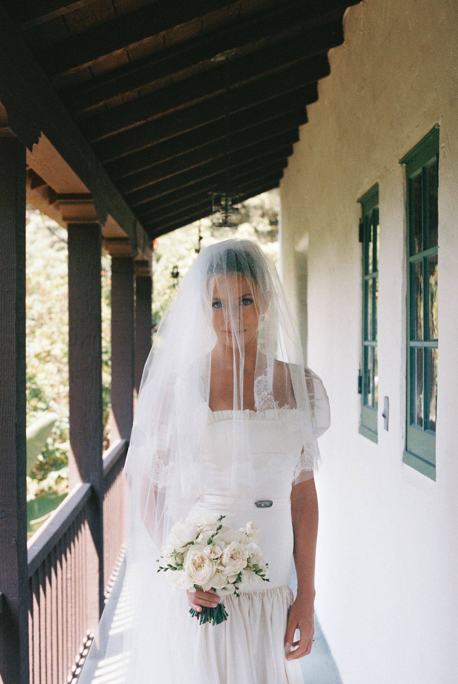 Long white bridal veils flowing in the breeze along a covered outdoor walkway with green shuttered windows.