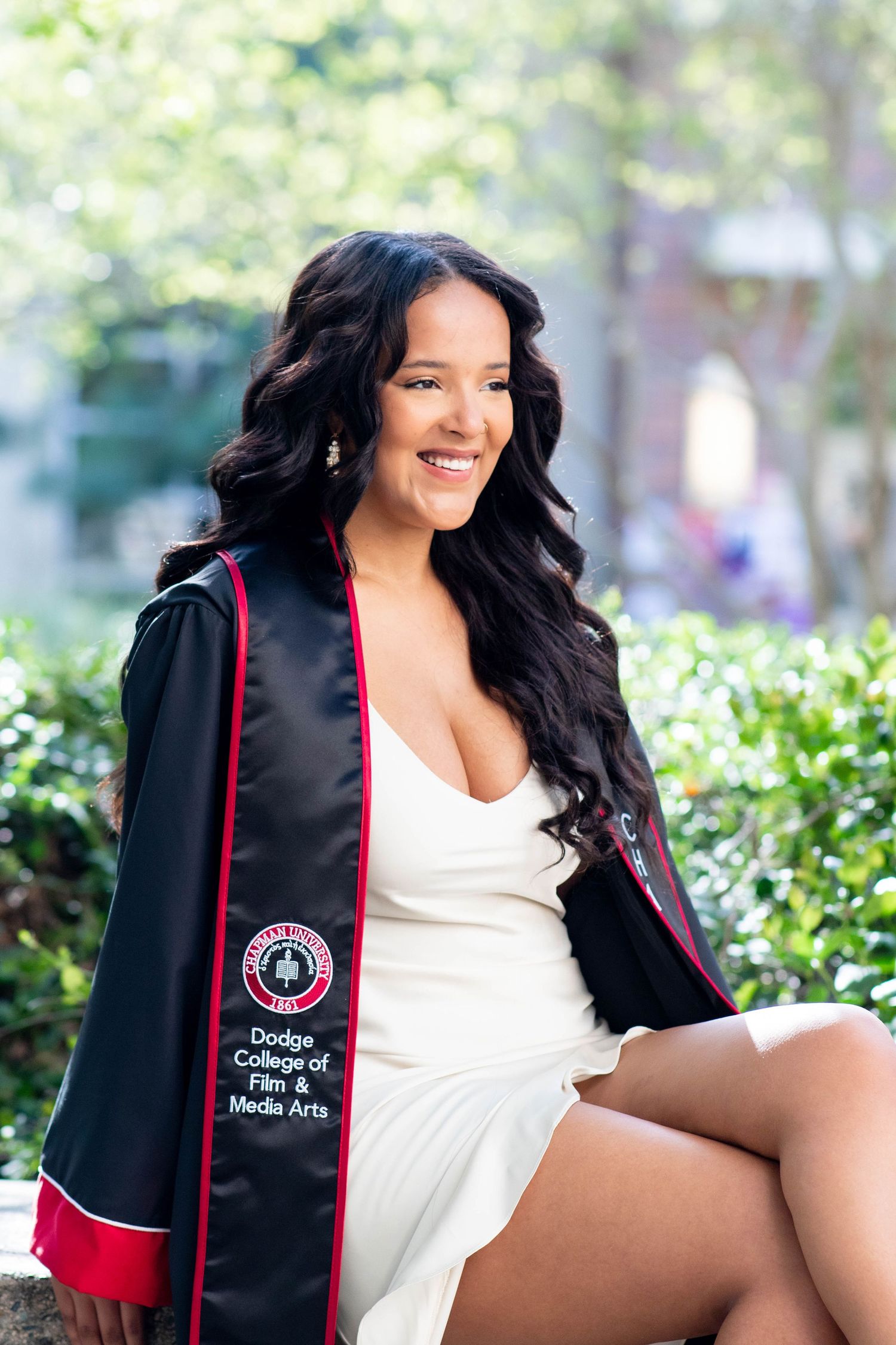 Graduate in black and red academic stole poses outdoors in white dress against blurred garden background.