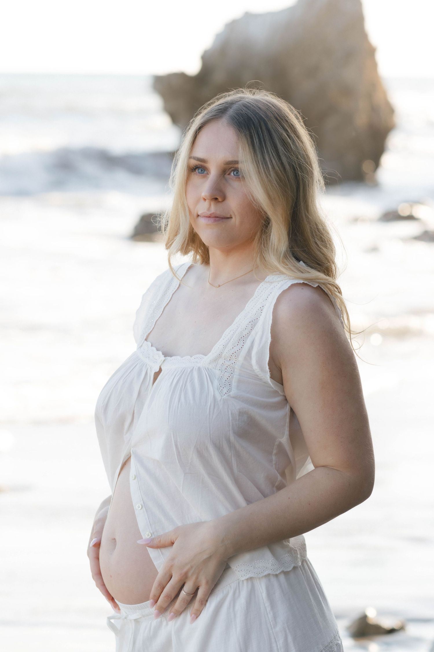 Pregnant woman in white dress poses on beach at sunset with glowing golden light in background.