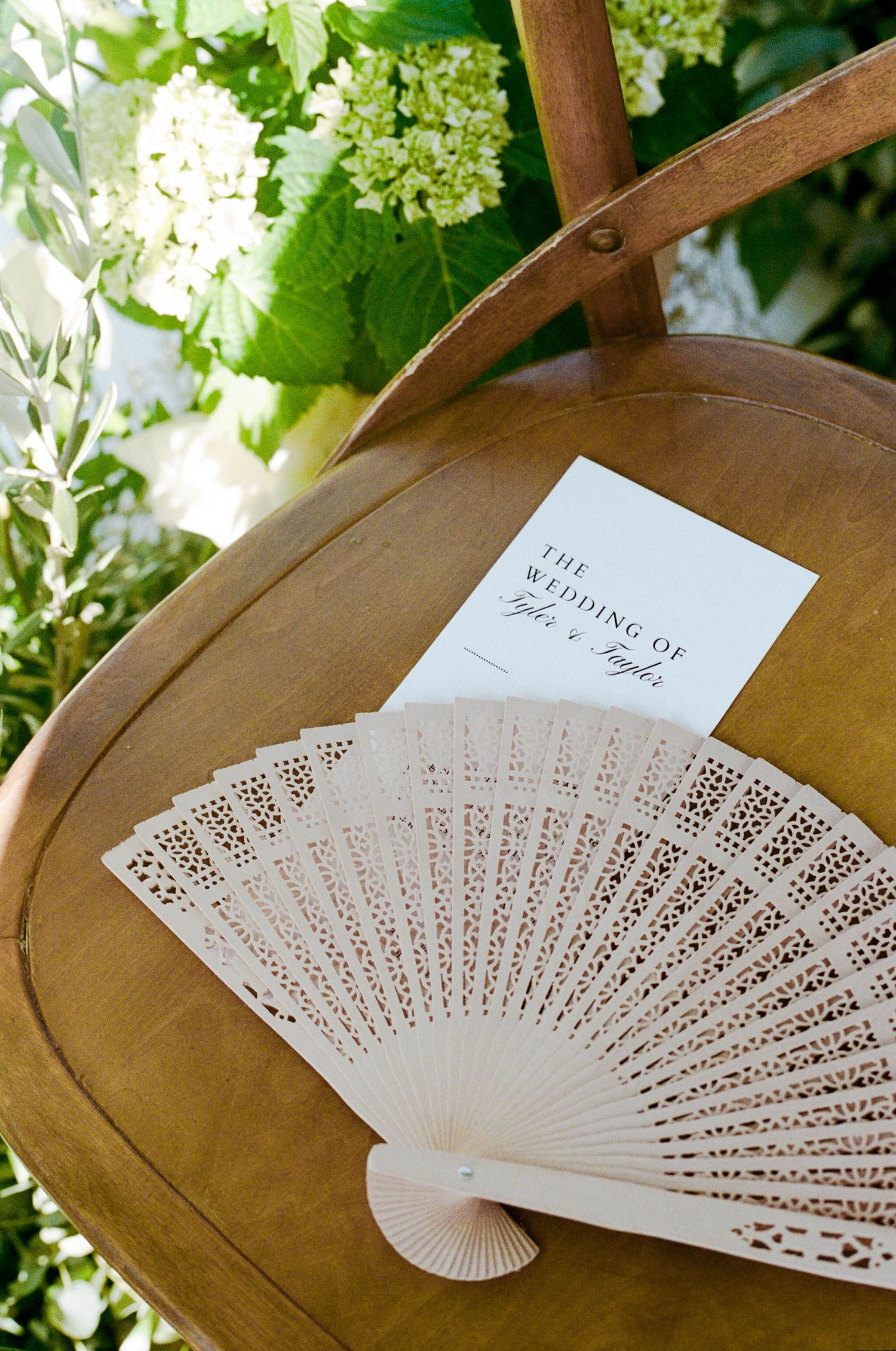 Decorative white paper fans and wedding programs displayed on wooden chairs at an outdoor venue.