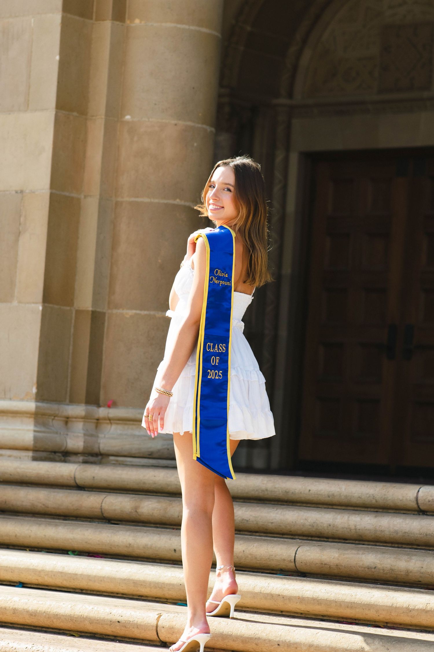 Graduate in white dress and blue sash poses on stone steps of campus building.